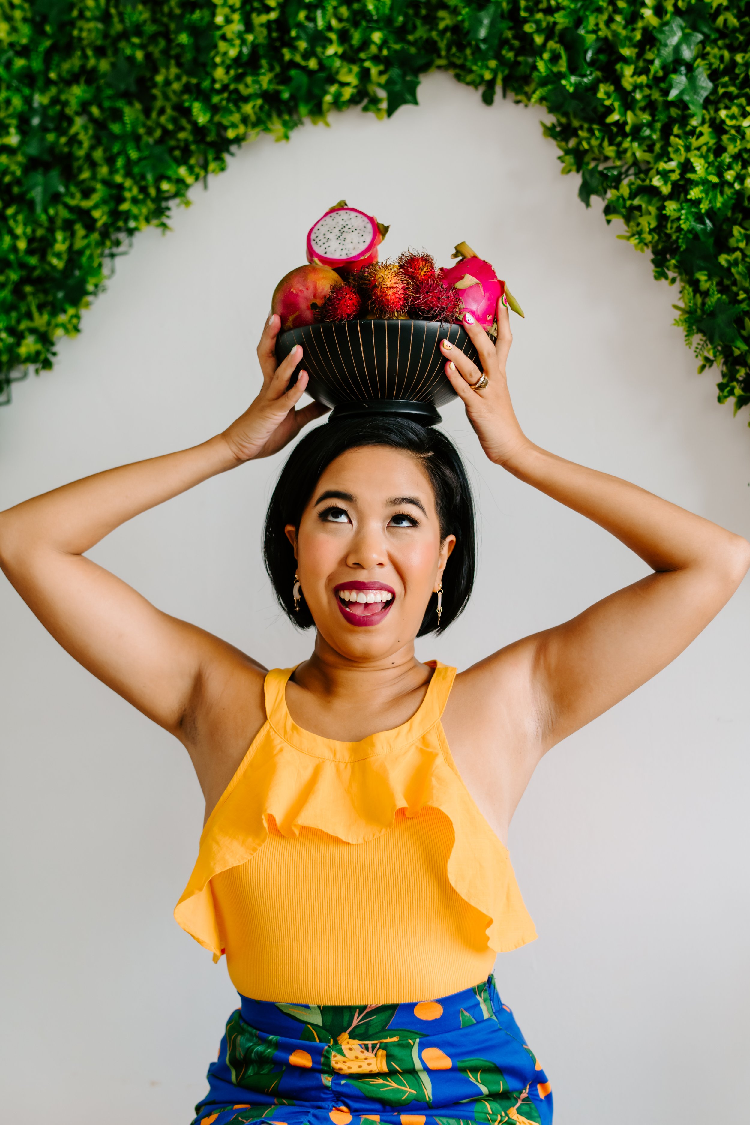 A woman with short dark hair smiling and wearing a yellow ruffled top and blue shorts with orange and green botanical print, holding a black bowl of tropical fruits on her head, against a white background with green foliage.