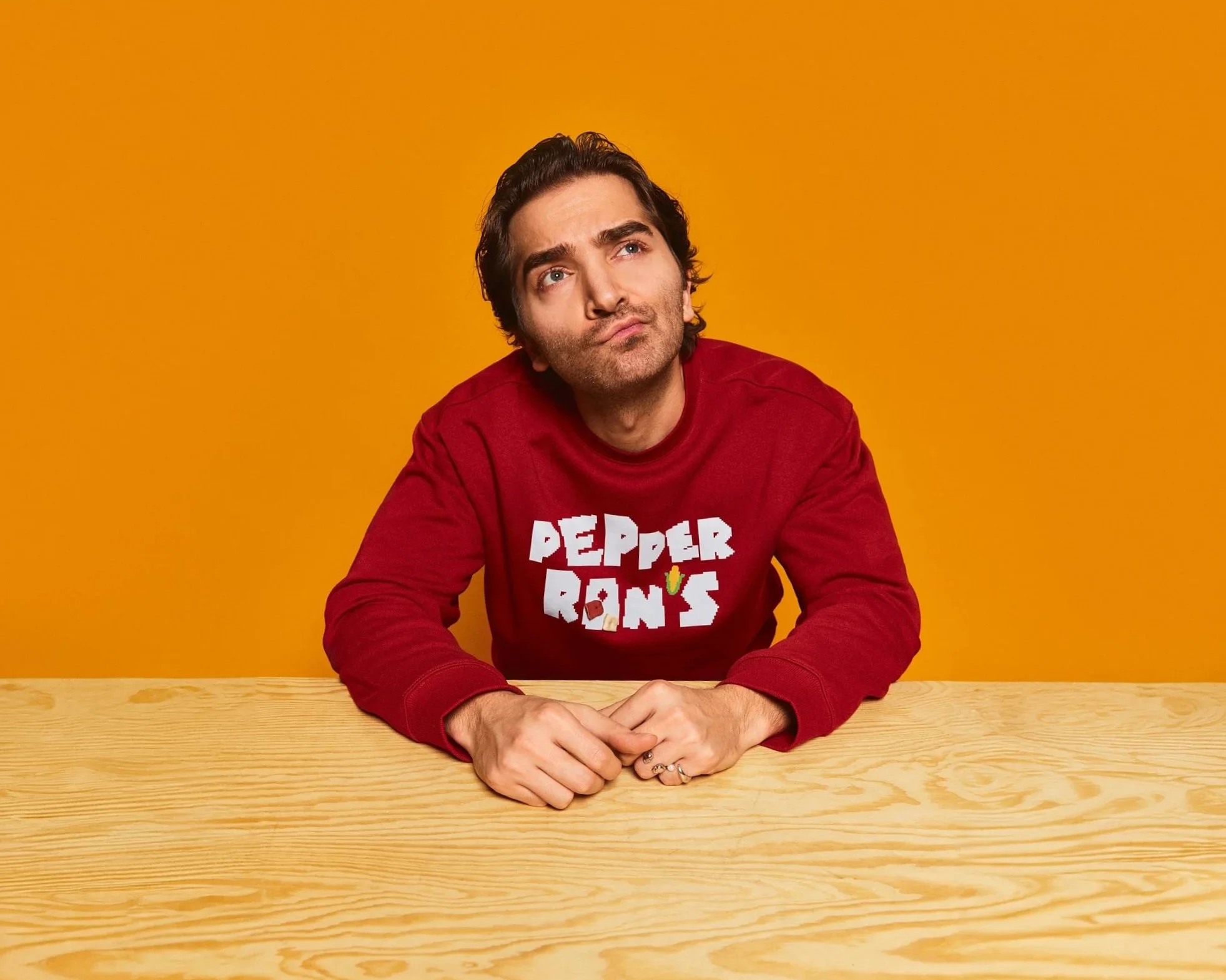 Person wearing a red "Pepper Ron's" sweatshirt sitting at a wooden table against an orange background, looking thoughtfully upward.