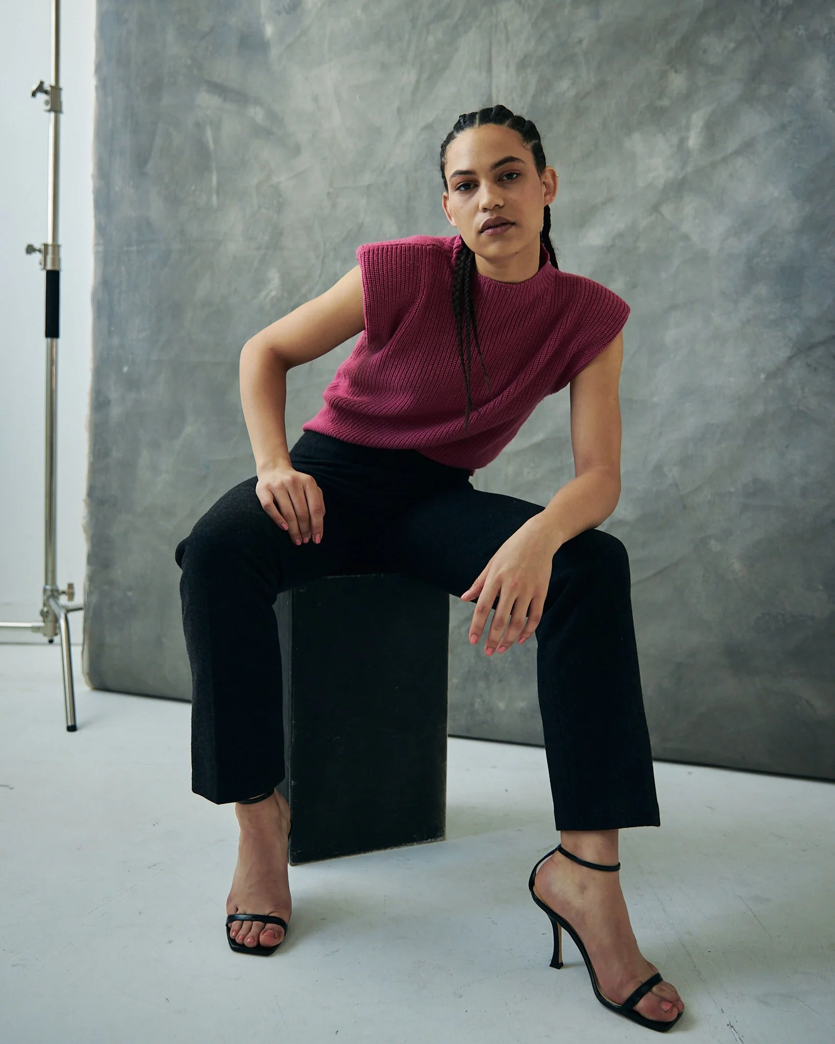 Person wearing a pink sleeveless top and black pants, sitting on a black cube against a textured gray background.