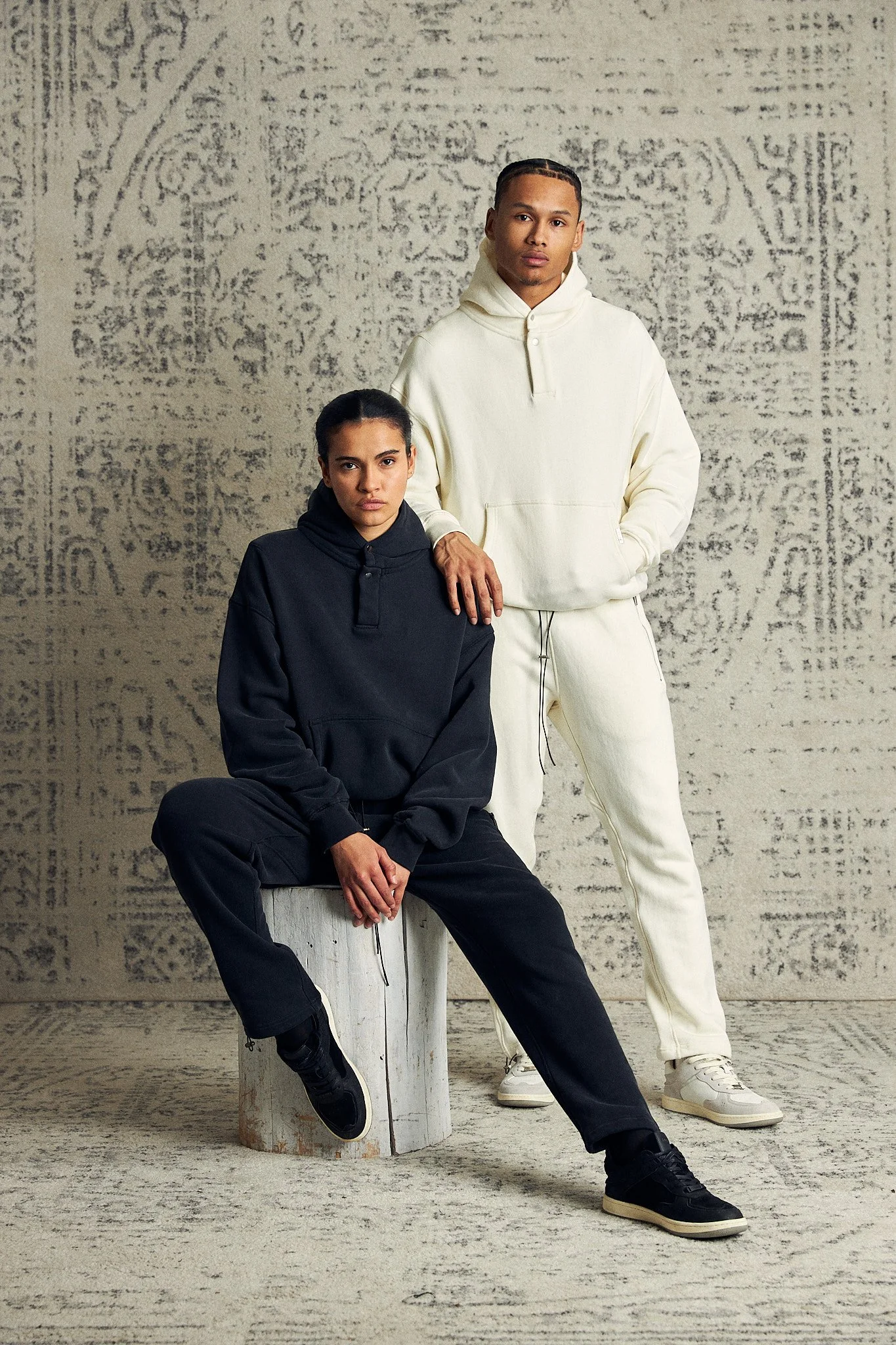 Two people wearing hoodies and sweatpants in a fashion photo shoot, with one sitting and the other standing against a textured background.