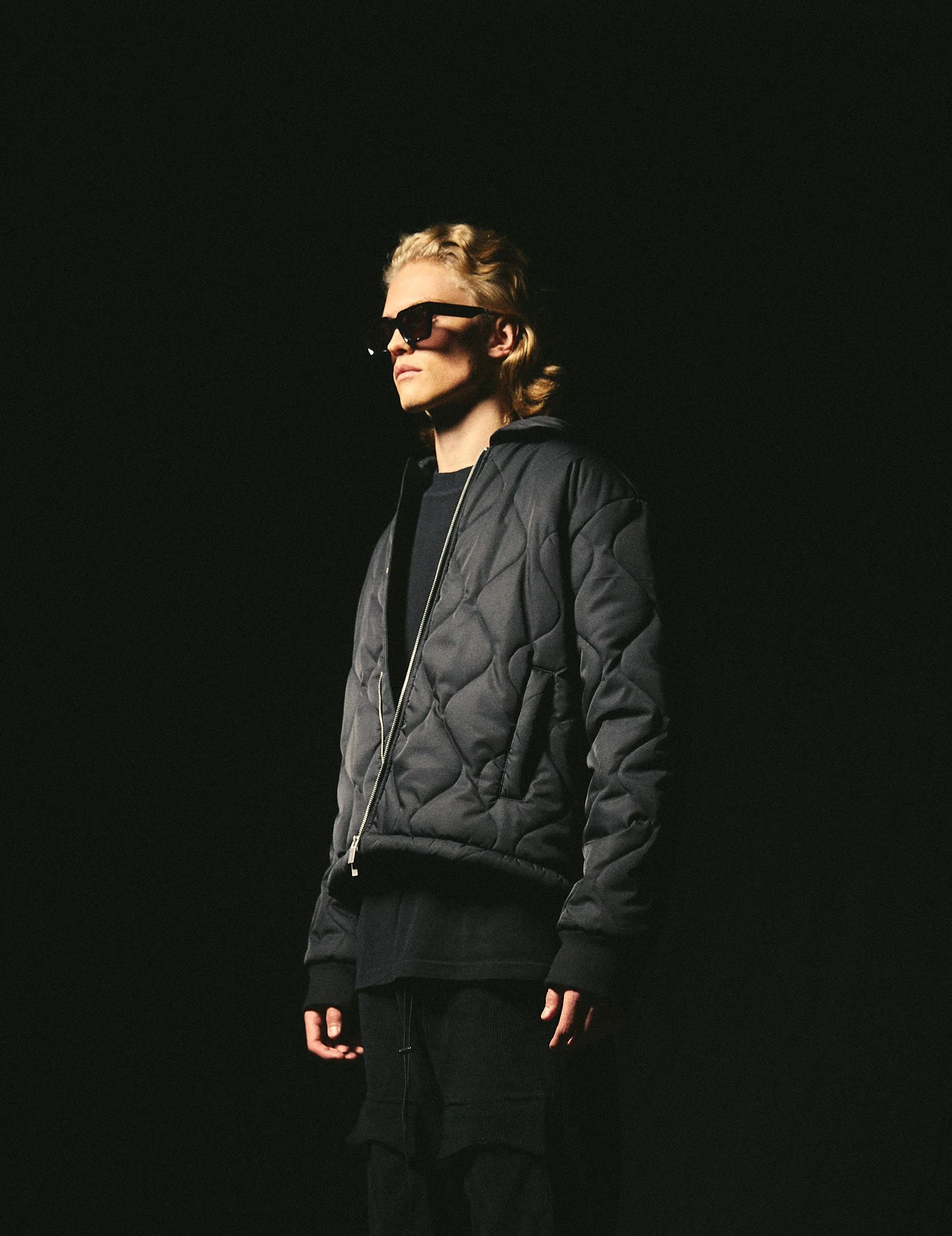 Person wearing sunglasses and a quilted jacket in dark setting