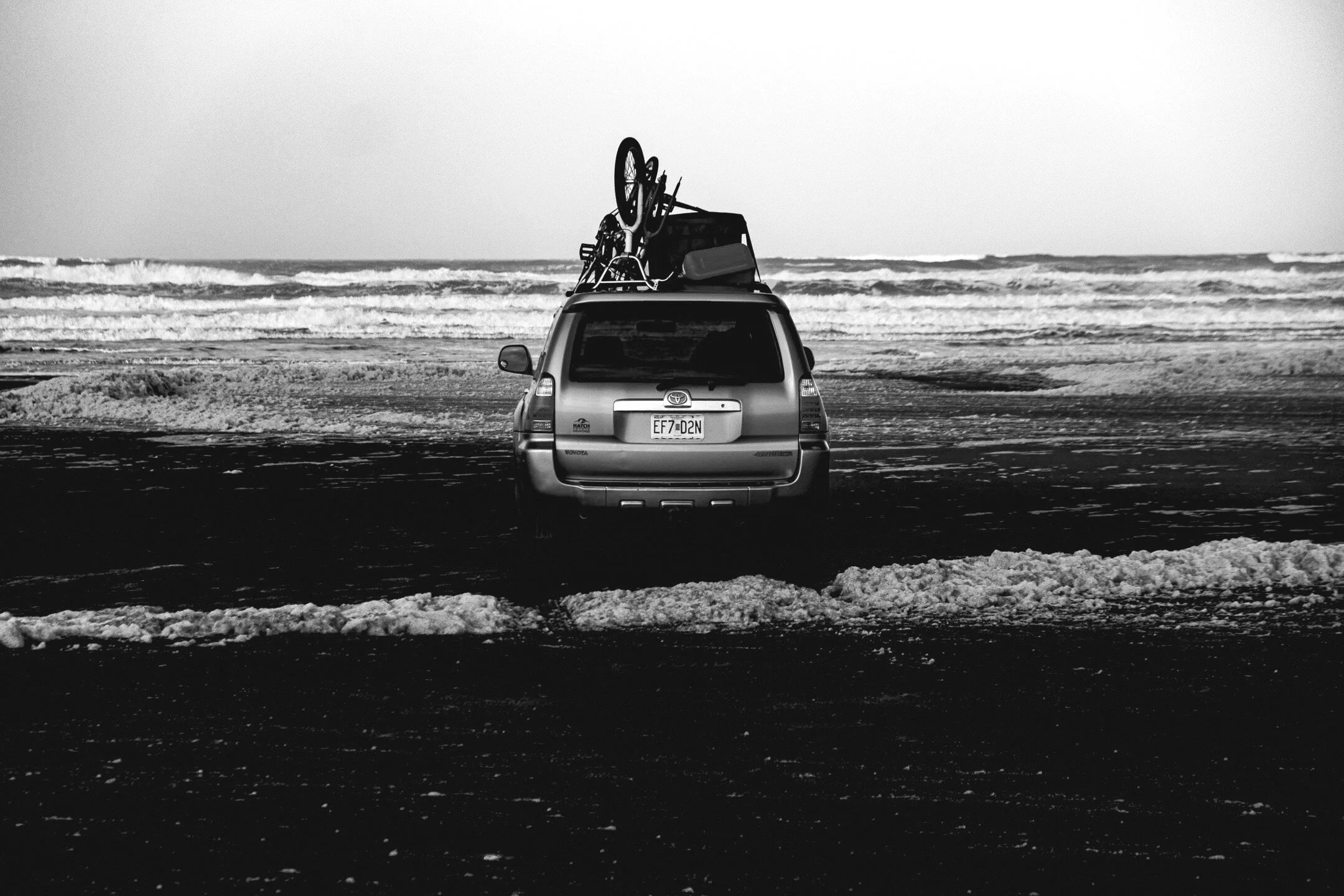 A black and white photo of a 4th generation Toyota 4runner SUV parked on a beach in front of crashing waves. Strapped to the hood area road bike, a BMX bike, a gas can, and a luggage tote.