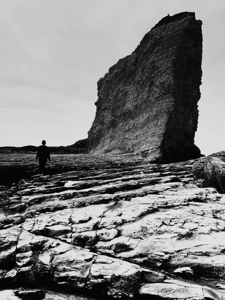 A small figure walks across a rock shelf beneath a sea stack