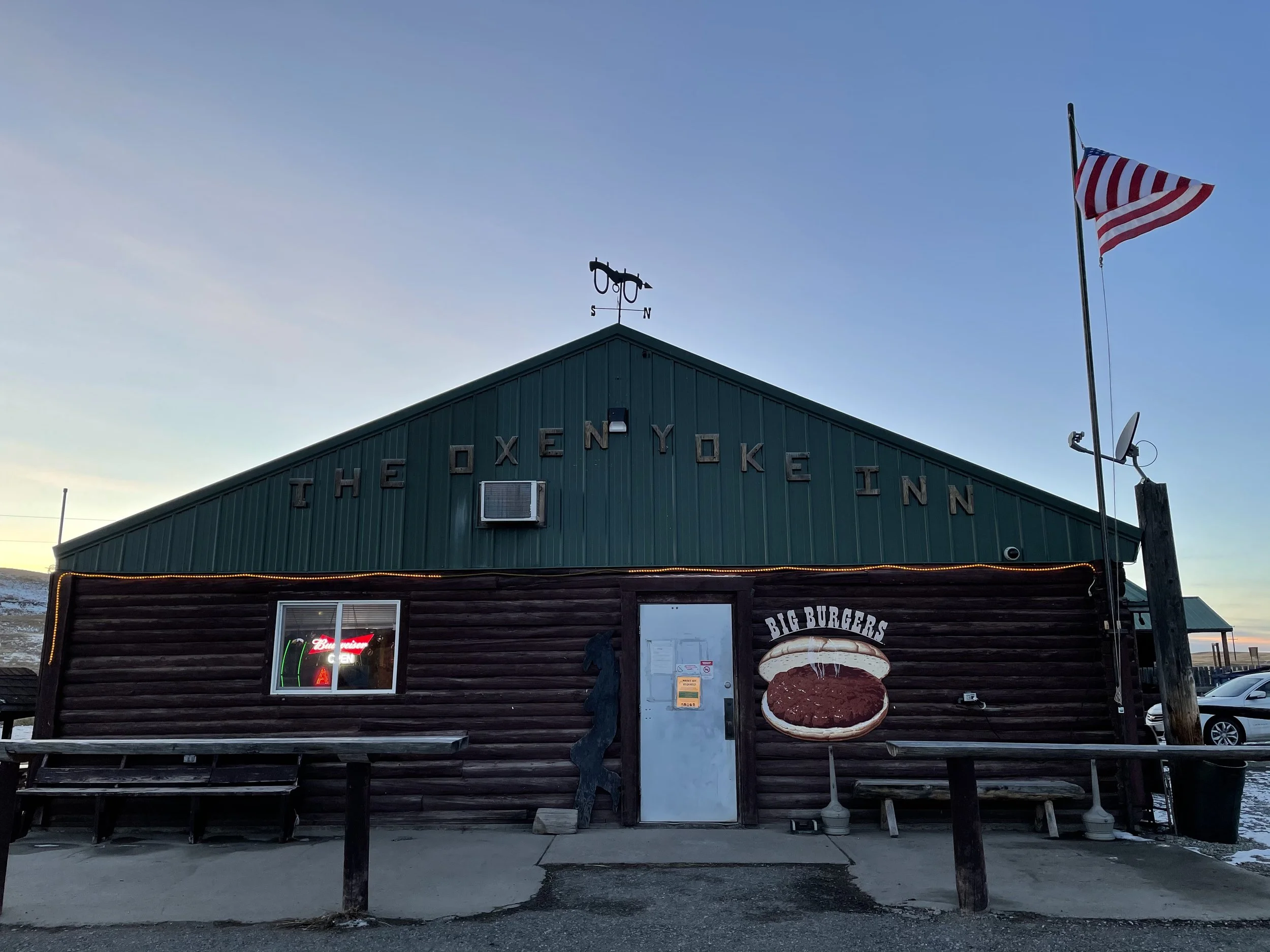 A rustic log and steel building with wooden letters reading The Oxen Yoke In. An American flag waves on a pole to the right. A giant hamburger sign is on the right of the entrance and reads BIG BURGERS