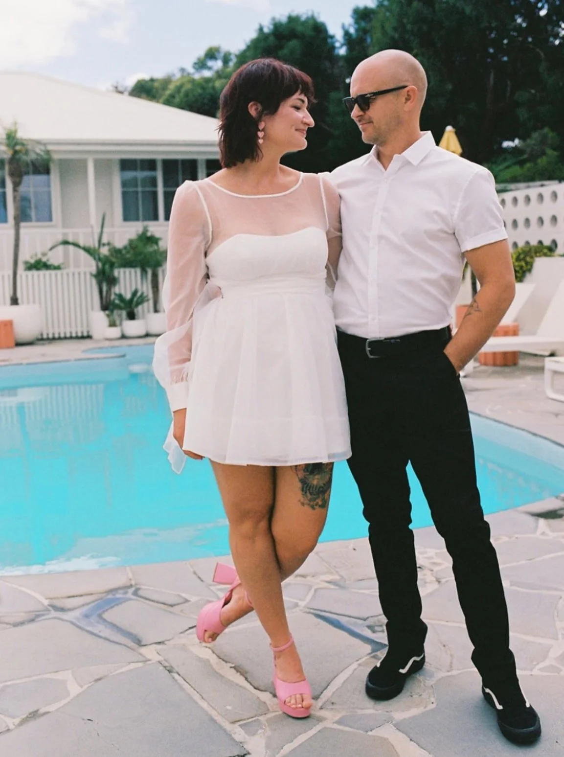 A woman and a man standing next to each other by a swimming pool, smiling and looking at each other. The woman is wearing a white dress, pink high heels, and pink earrings. The man is dressed in a white shirt, black pants, and sunglasses.