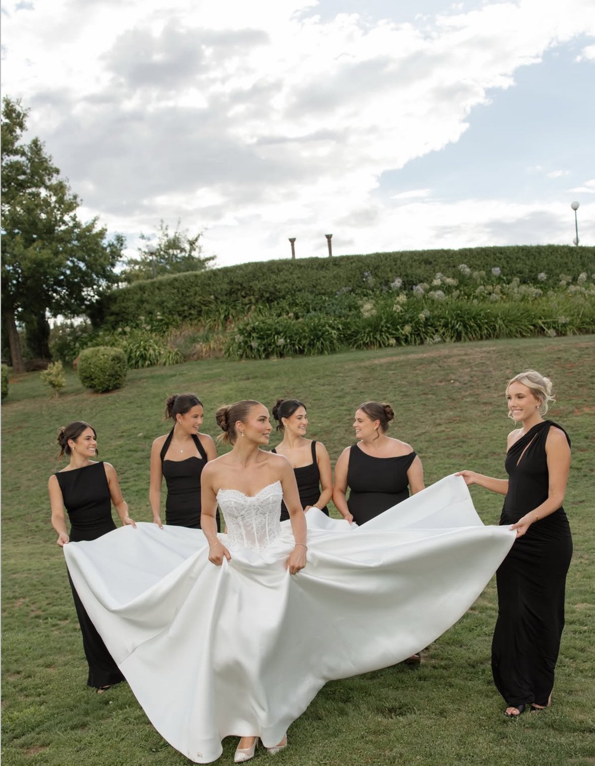 A bride in a white wedding gown and five bridesmaids in black dresses outdoors in a grassy area with trees and a cloudy sky.