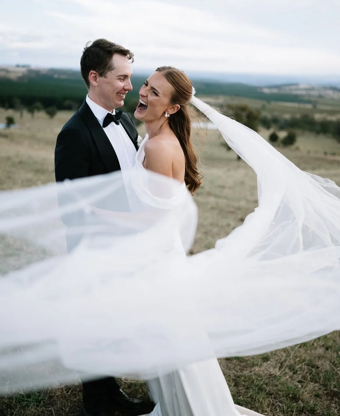 A bride and groom smiling and laughing outdoors on a wedding day, with the bride's veil flowing in the wind, set in a rural landscape.