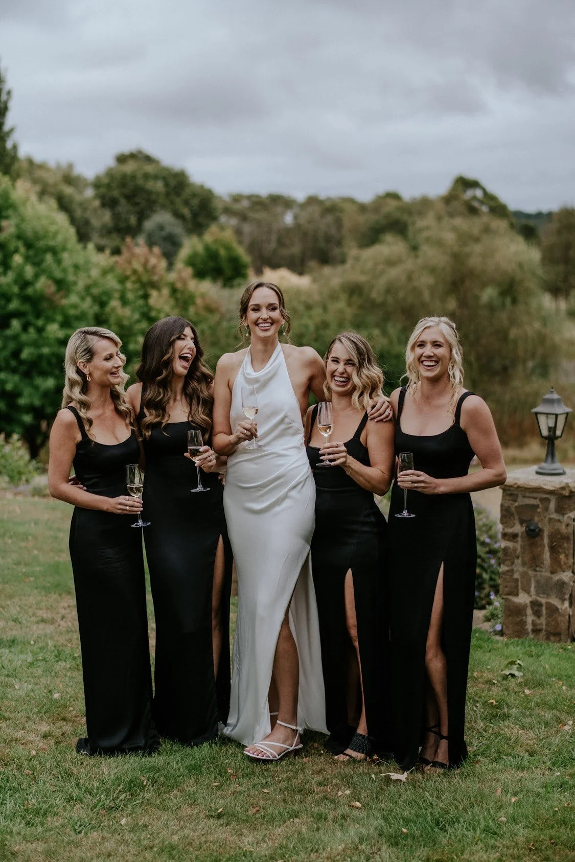 Group of five women in formal attire, holding glasses of champagne outdoors, smiling and enjoying an event. Four women are wearing matching black dresses with high slits, while the woman in the center wears a white dress. The background features trees and a cloudy sky.