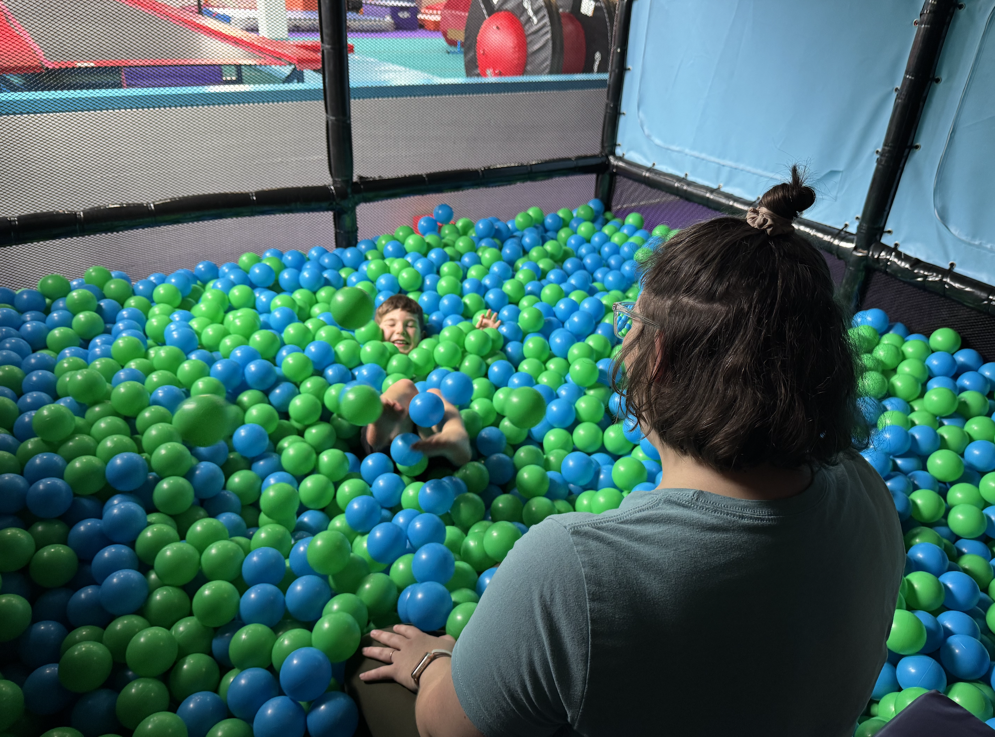 A young boy smiles as he falls into a ball pit full of blue and green balls with only his face and feet showing. A woman with short brown hair wearing a blue shirt watches him with her back to the camera.