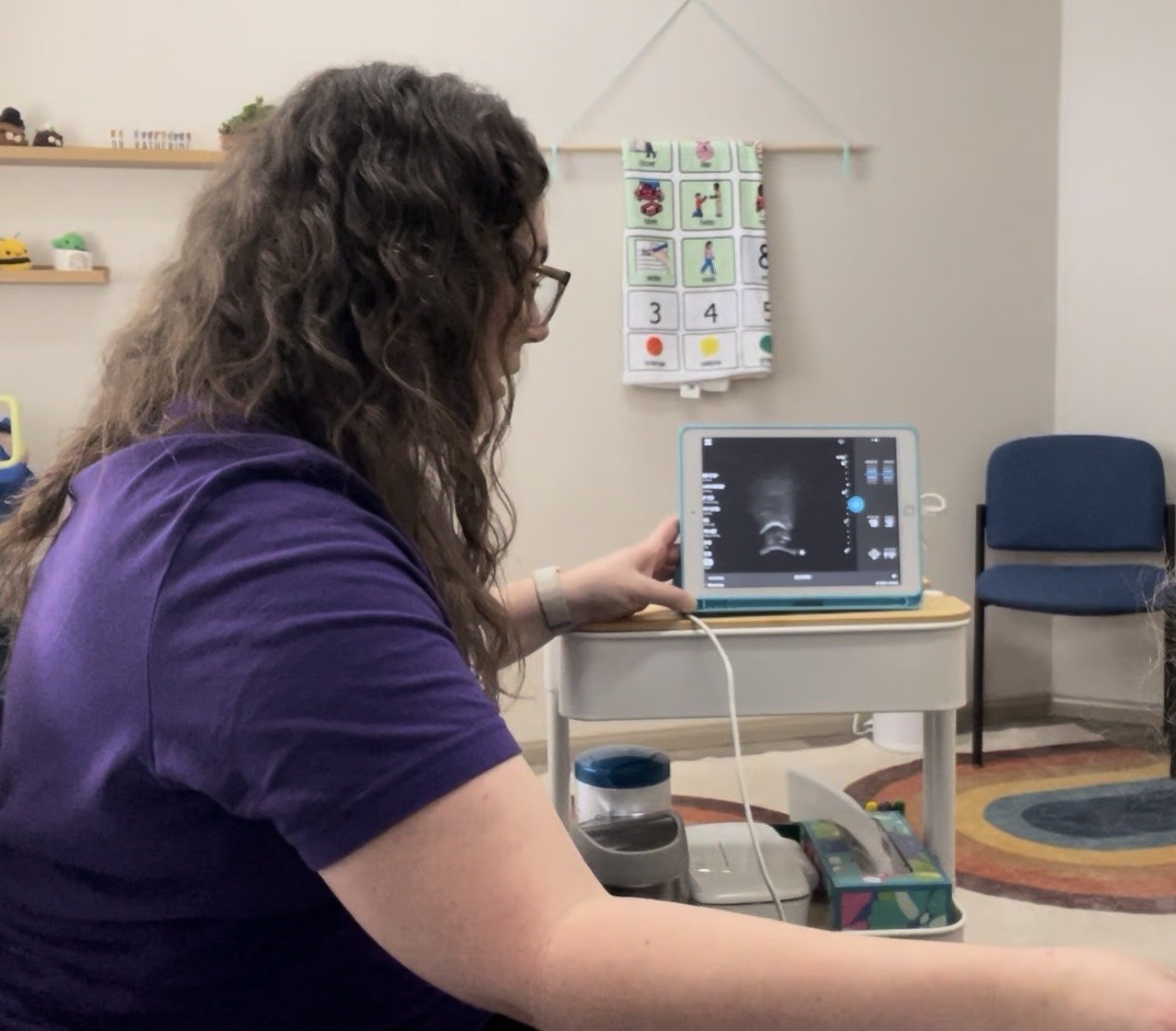 A woman with long, wavy, brown hair and a purple shirt holds an iPad with an ultrasound image of the tongue.