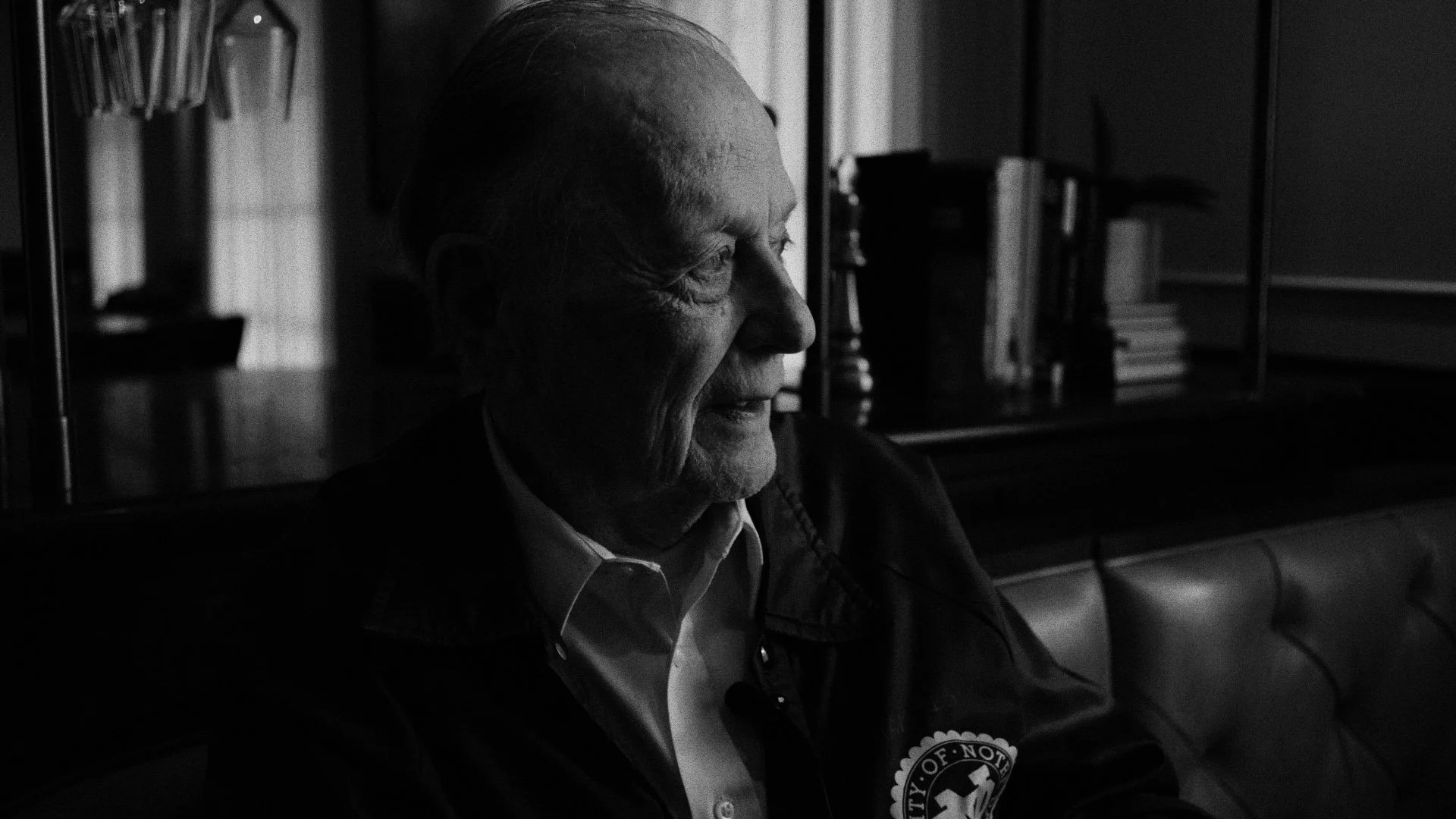 A black and white photo of an elderly man with a passionate, smiling expression, sitting indoors with a bookshelf and books in the background.