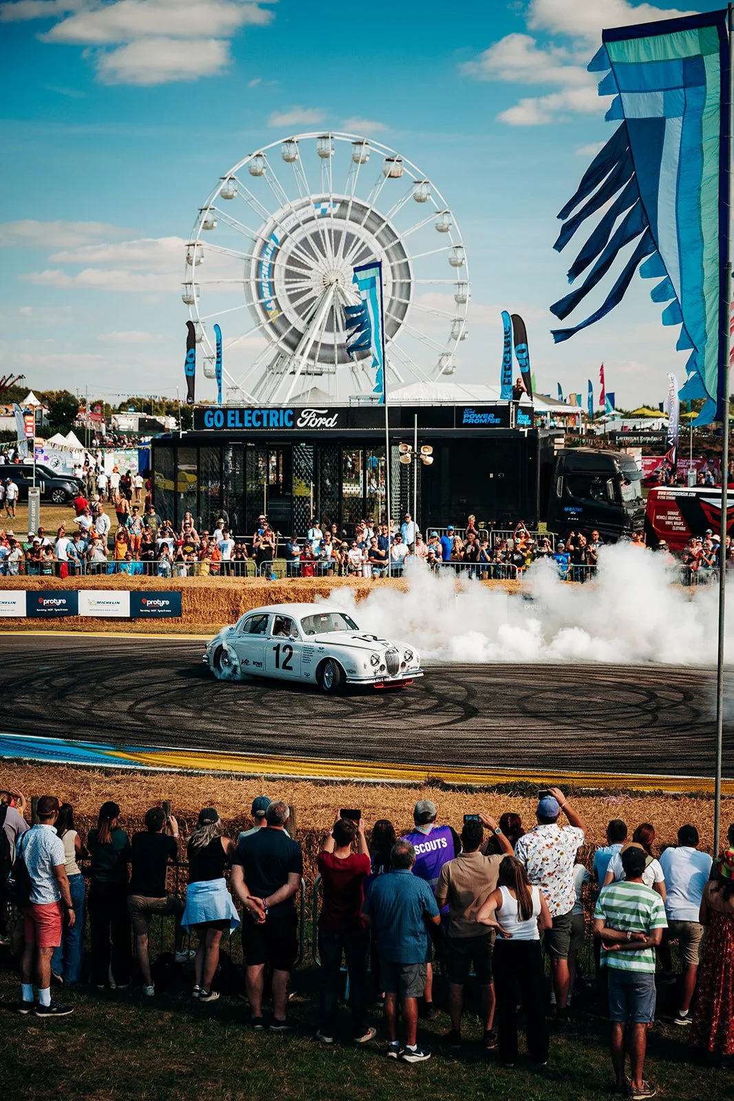 Classic white race car spinning in a cloud of tyre smoke at CarFest, with a crowd and Ferris wheel behind