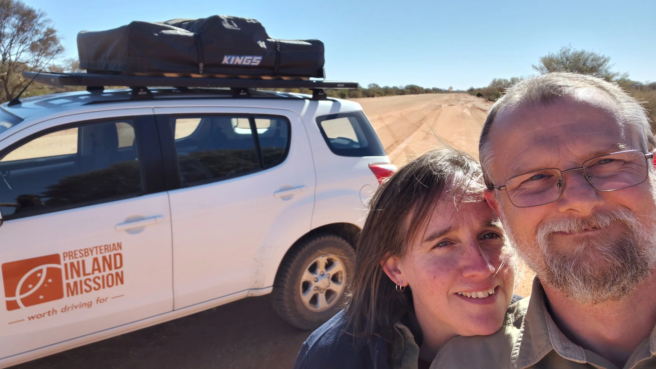 Two people taking a selfie in front of a white vehicle with 'Presbyterian Inland Mission' logo, on a dusty, rural road surrounded by dry bushes and clear blue sky.