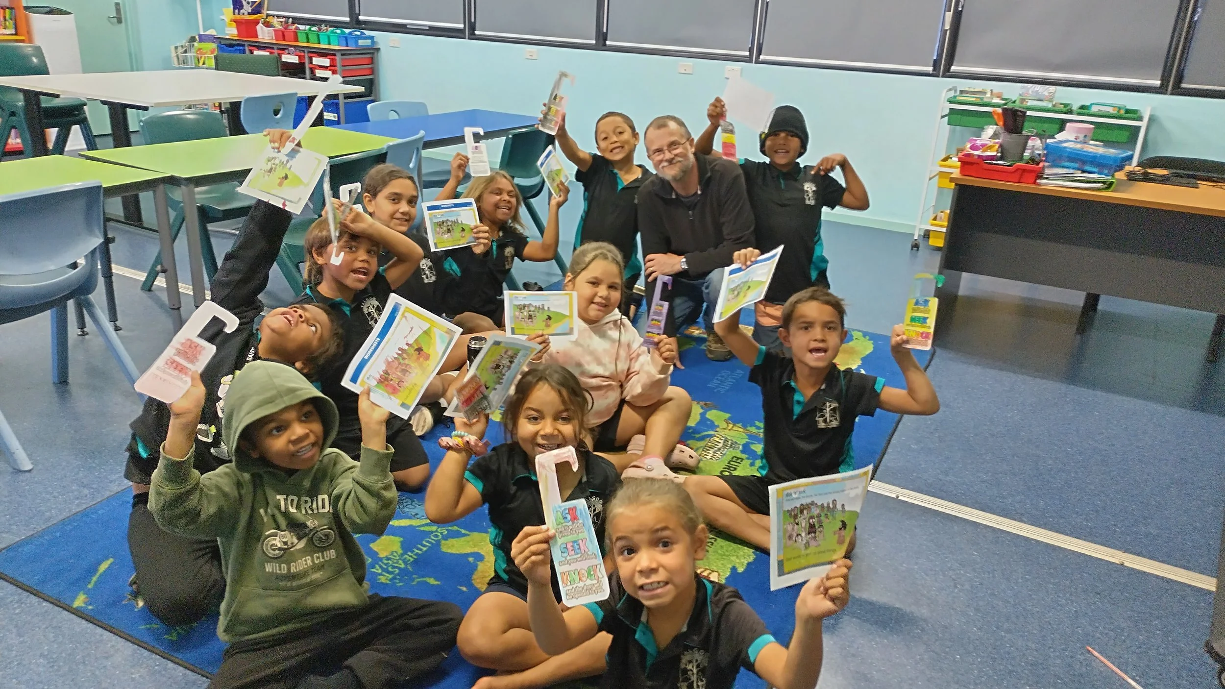 A group of children and an adult in a classroom, holding up books or pamphlets, smiling and celebrating.