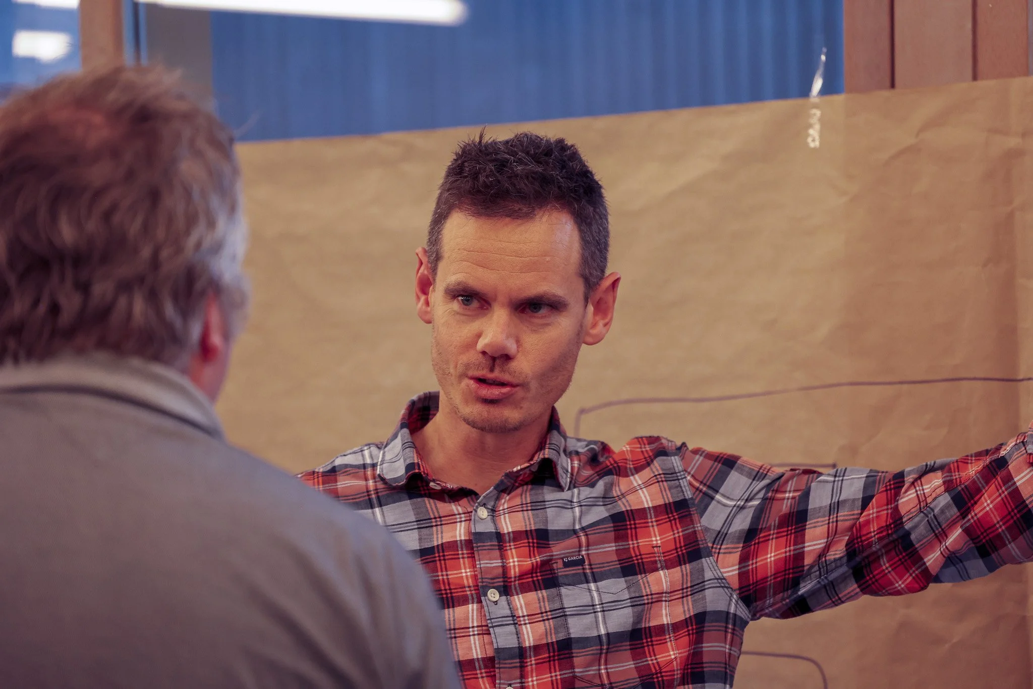 Two men having a conversation in an office or meeting room. One man is facing away from the camera, and the other man is facing the camera, wearing a plaid shirt and gesturing with his arm. The background features brown paper and wooden decor.