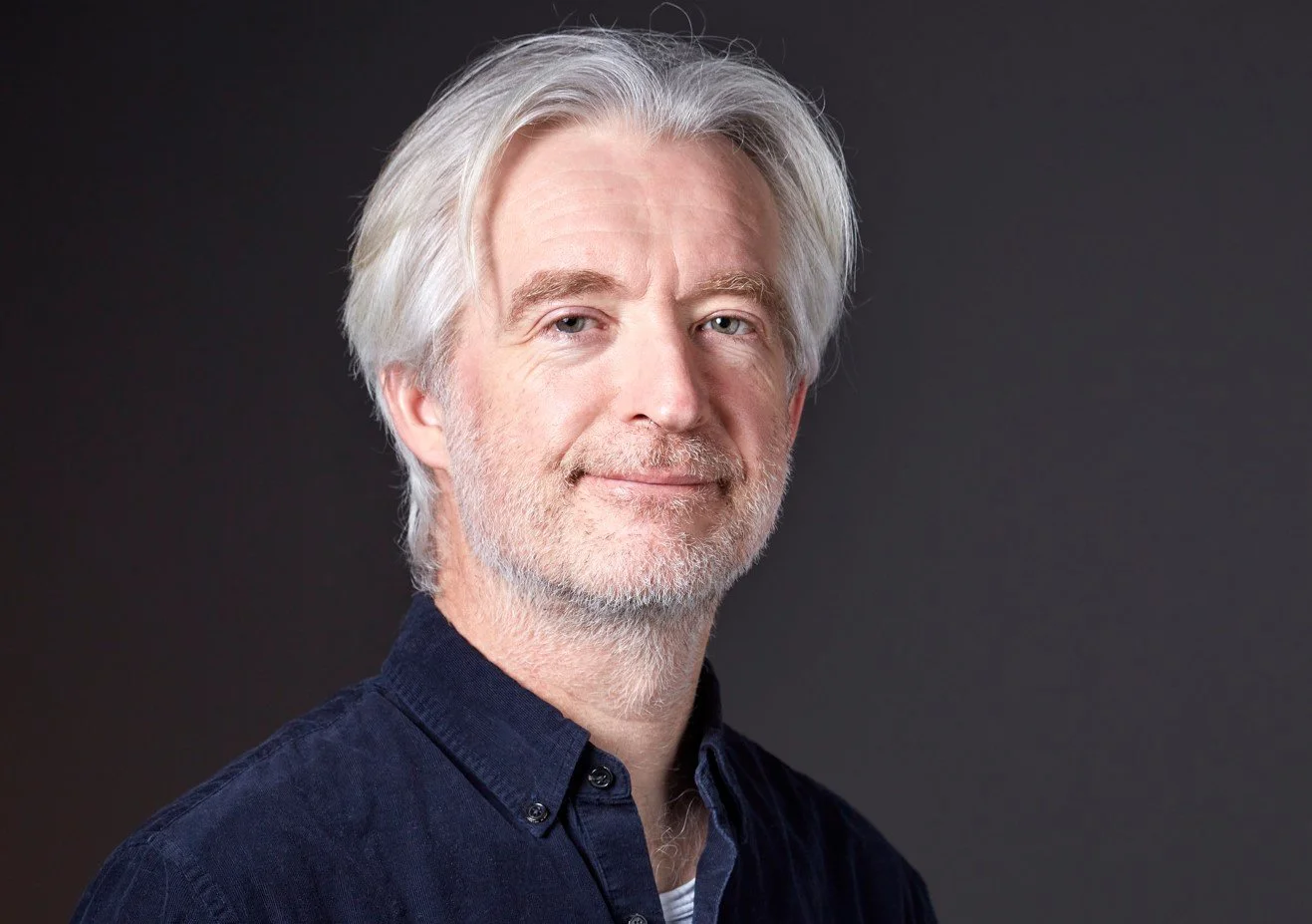 A middle-aged man with white hair and a beard, wearing a dark blue shirt, looking at the camera with a slight smile against a dark background.