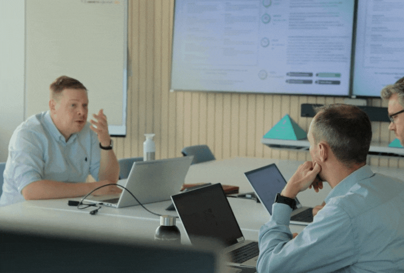 Three men in a meeting room with laptops, a large screen displaying data behind them.