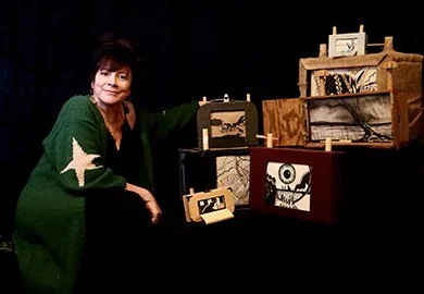 A woman with short hair wearing a green sweater with white stars, sitting beside a display of small wooden shadow boxes with various decorations, including an owl, a house, and abstract art, on a black background.