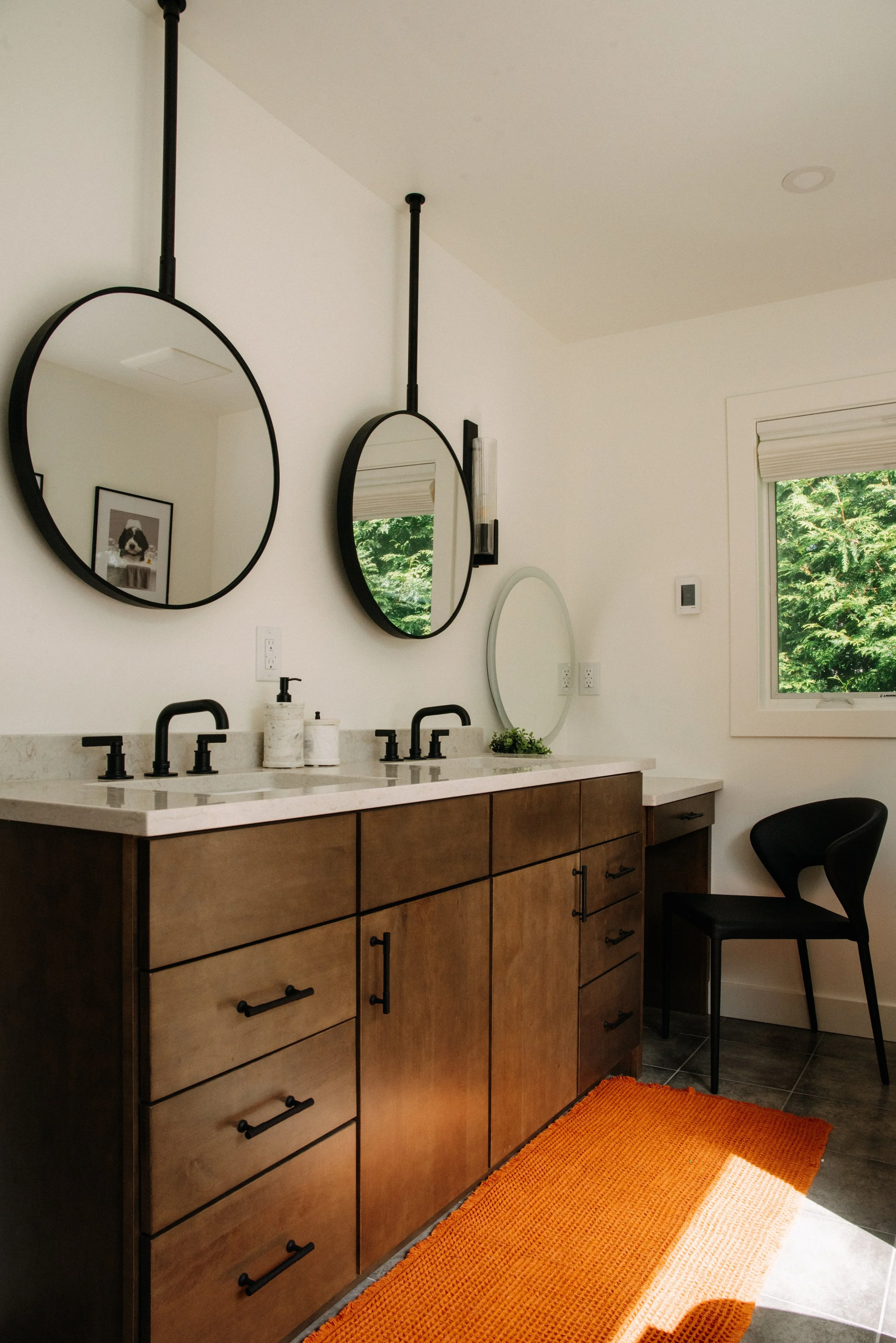 A modern bathroom with a double vanity featuring wood cabinets, white countertop, and black faucets. Two round mirrors are hung above the sinks, and a smaller oval mirror is on the wall. There are black light fixtures, a window showing green trees ou