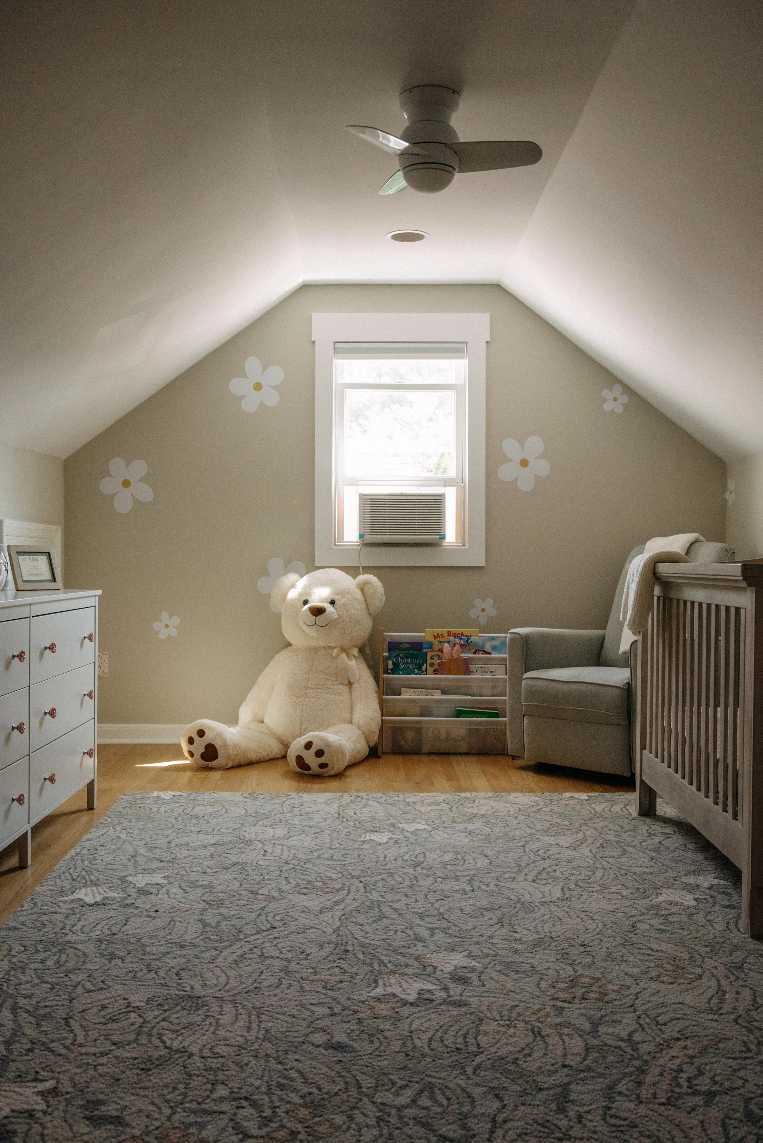A cozy child's bedroom with a large teddy bear sitting on the floor next to a plastic storage container filled with books, a gray armchair, and a white dresser. The room has a window with an air conditioning unit, angled ceilings, a ceiling fan, and