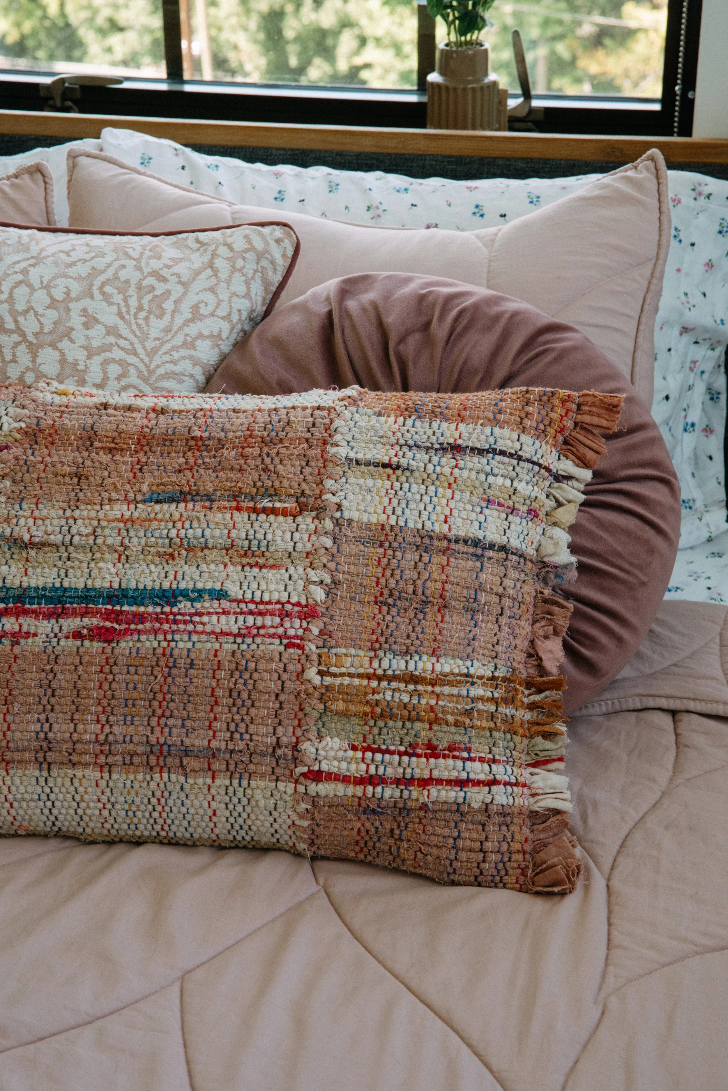 A bed with a beige quilt, decorated with multiple pillows including a large textured pillow, a floral pillow, and a plain cushion, set near a window with a potted plant on the sill.