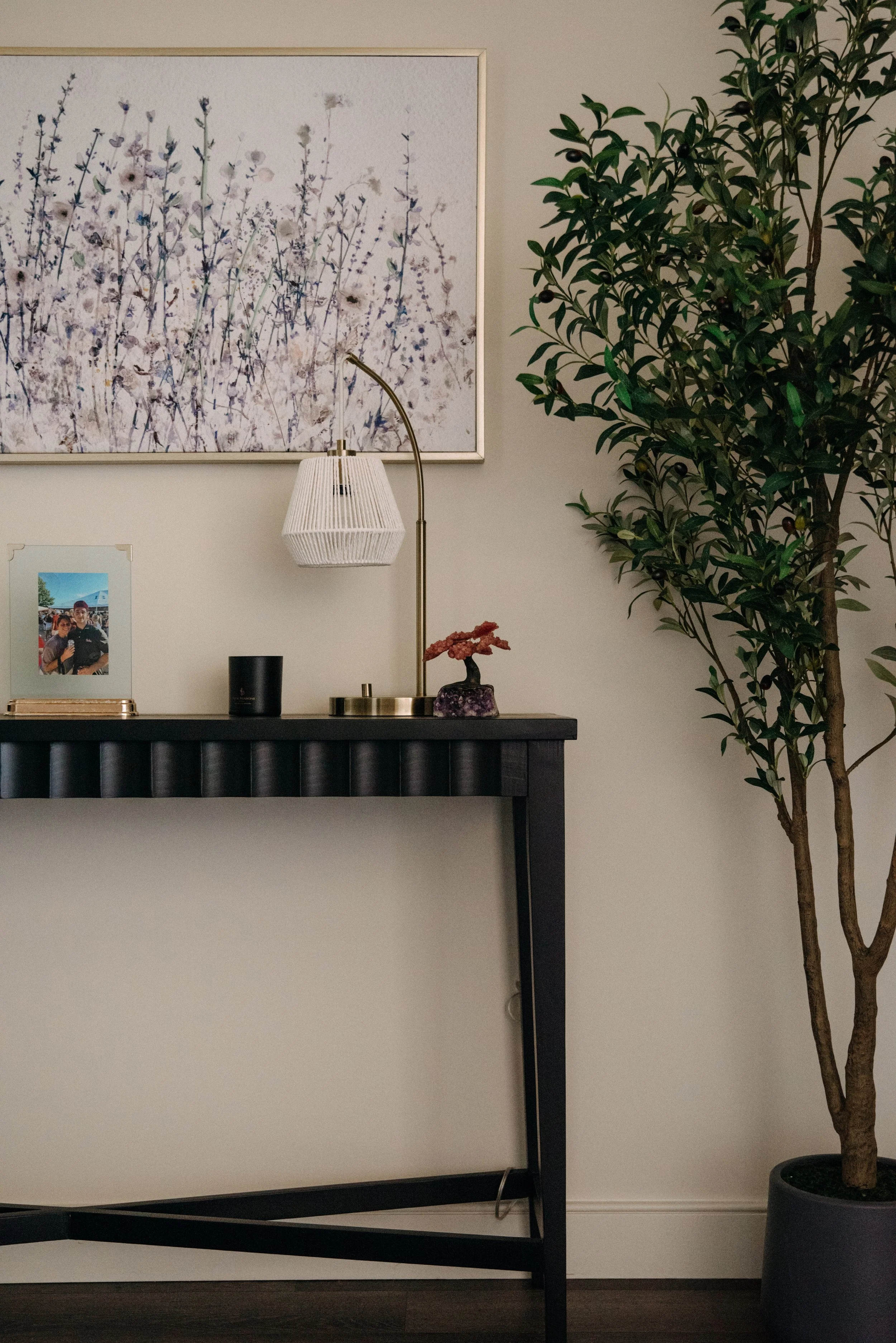 A black console table with decorative items including a framed photo, a black speaker, a gold disc candle holder, a pink coral-like sculpture, and a table lamp with a white shade, against a light-colored wall with a large floral painting and a tall p