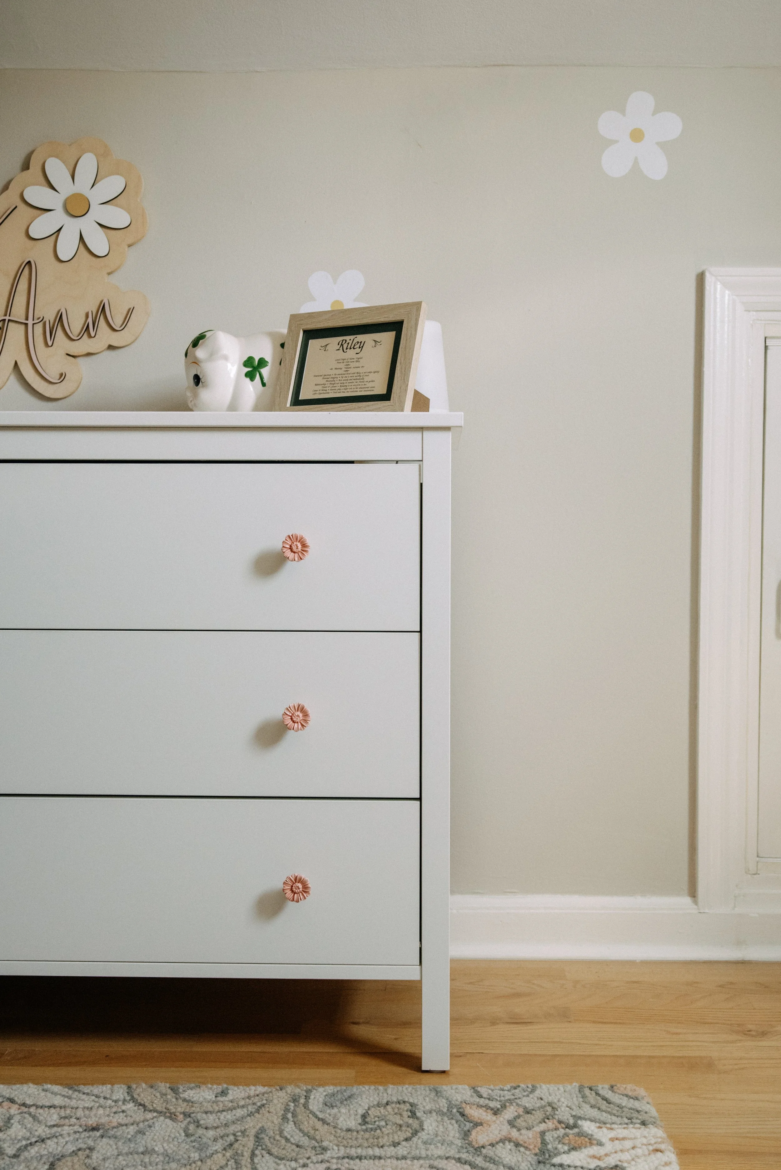 A white dresser with three drawers, each with pink flower-shaped handles, positioned on a light wood floor with a patterned rug in front. On top of the dresser are decorative items including a framed picture, a white ceramic animal, and a wooden sign