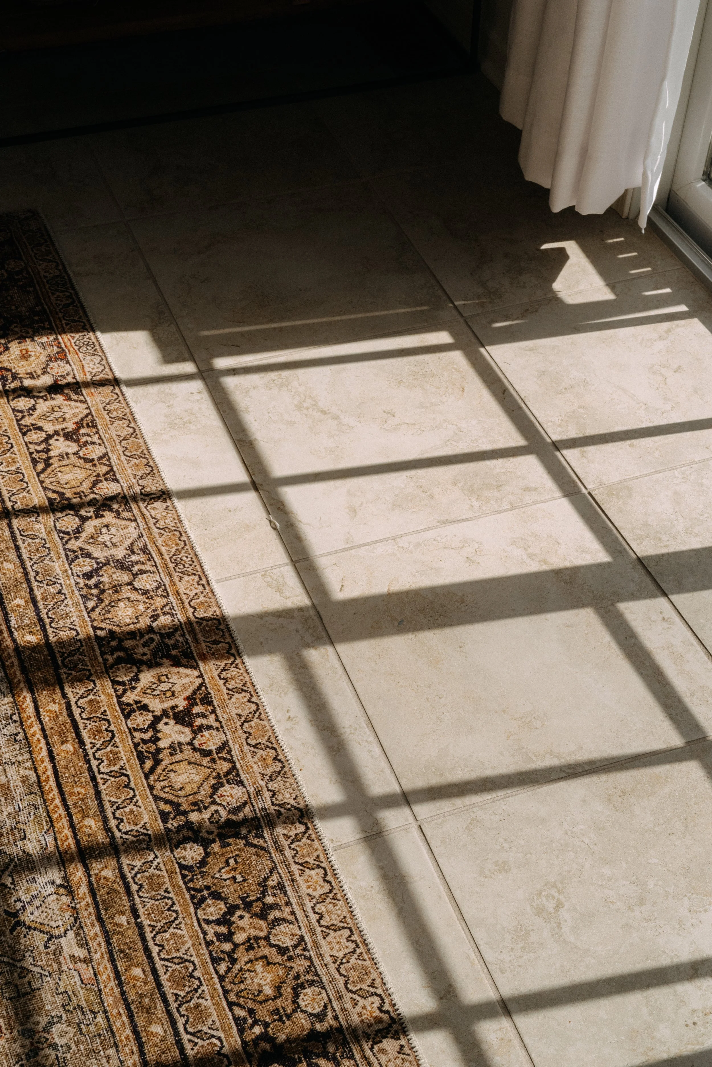 Sunlight casting shadows through a window onto a tiled floor and a patterned rug.