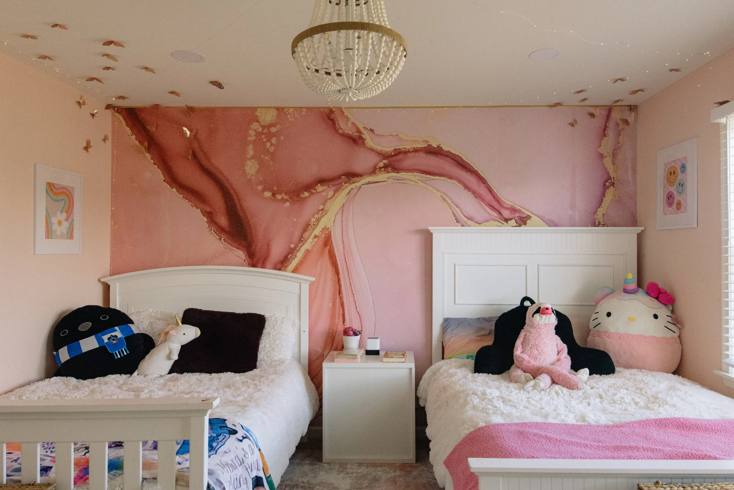 Bedroom with two white beds and plush toys, pink and gold marbled accent wall, ceiling with gold butterflies and a white chandelier, framed artwork with colorful smiley faces and abstract rainbow on walls, sunlight coming through window with white bl