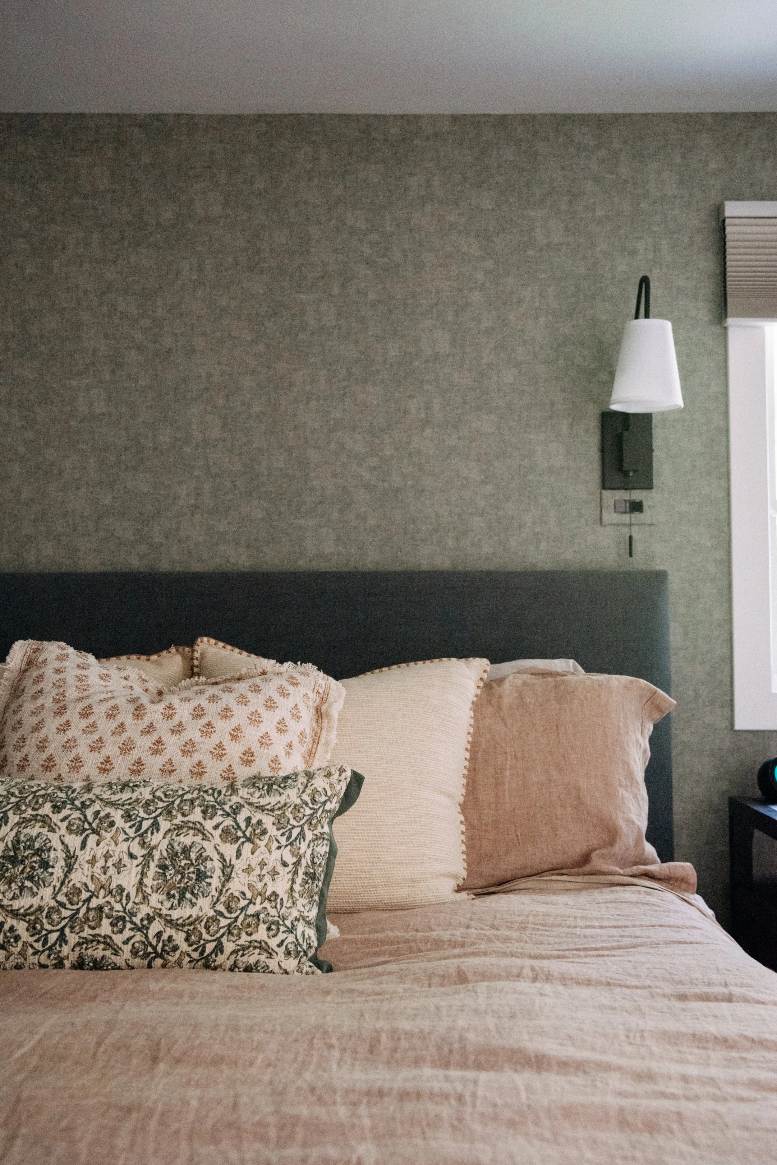 A bed with multiple pillows against a dark headboard in a bedroom with gray patterned wall, a wall-mounted light, and a small window.