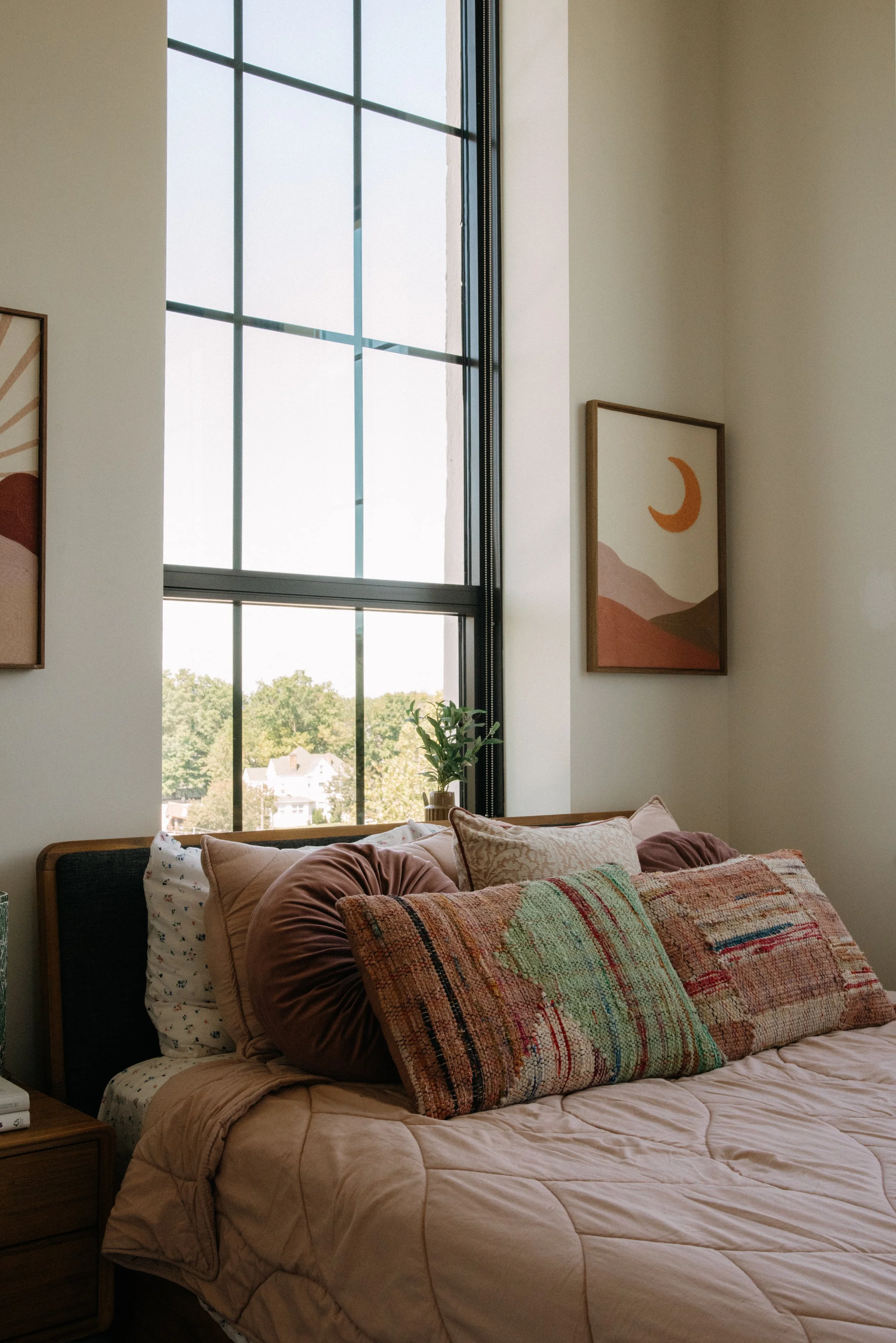 A cozy bedroom with a large window, decorated with framed artwork and a bed with multiple pillows and a beige quilt.