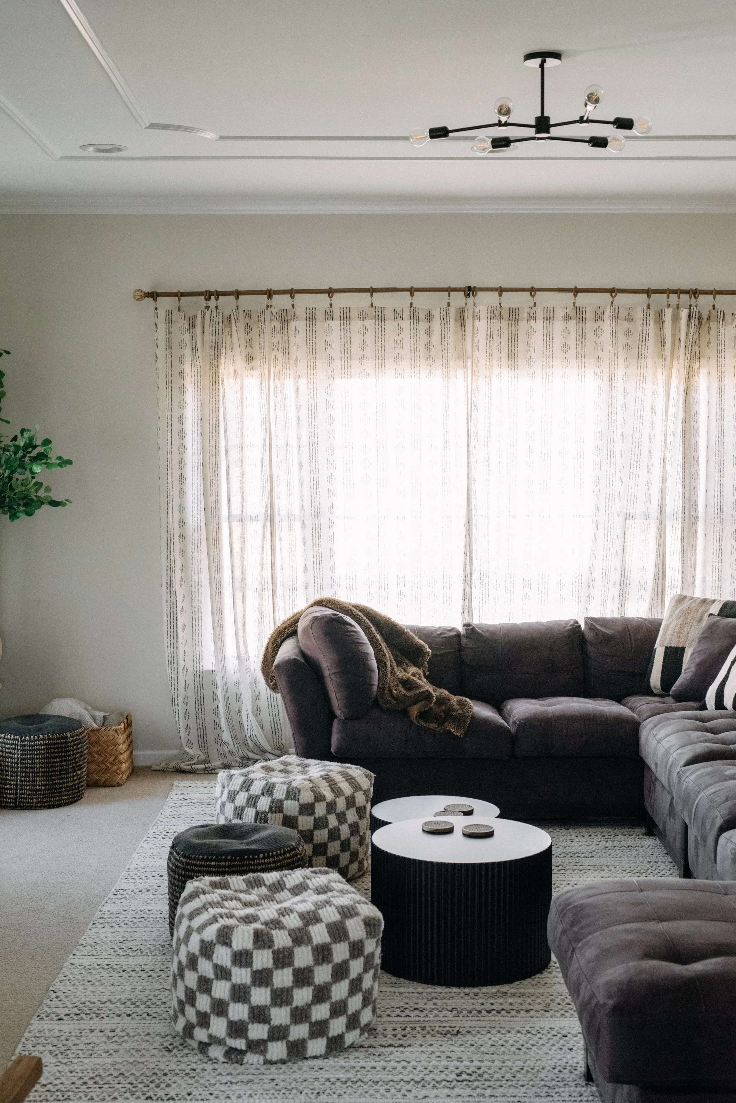 Living room with dark sectional sofa, checkered ottomans, round black coffee tables, beige curtains, and a modern black chandelier.
