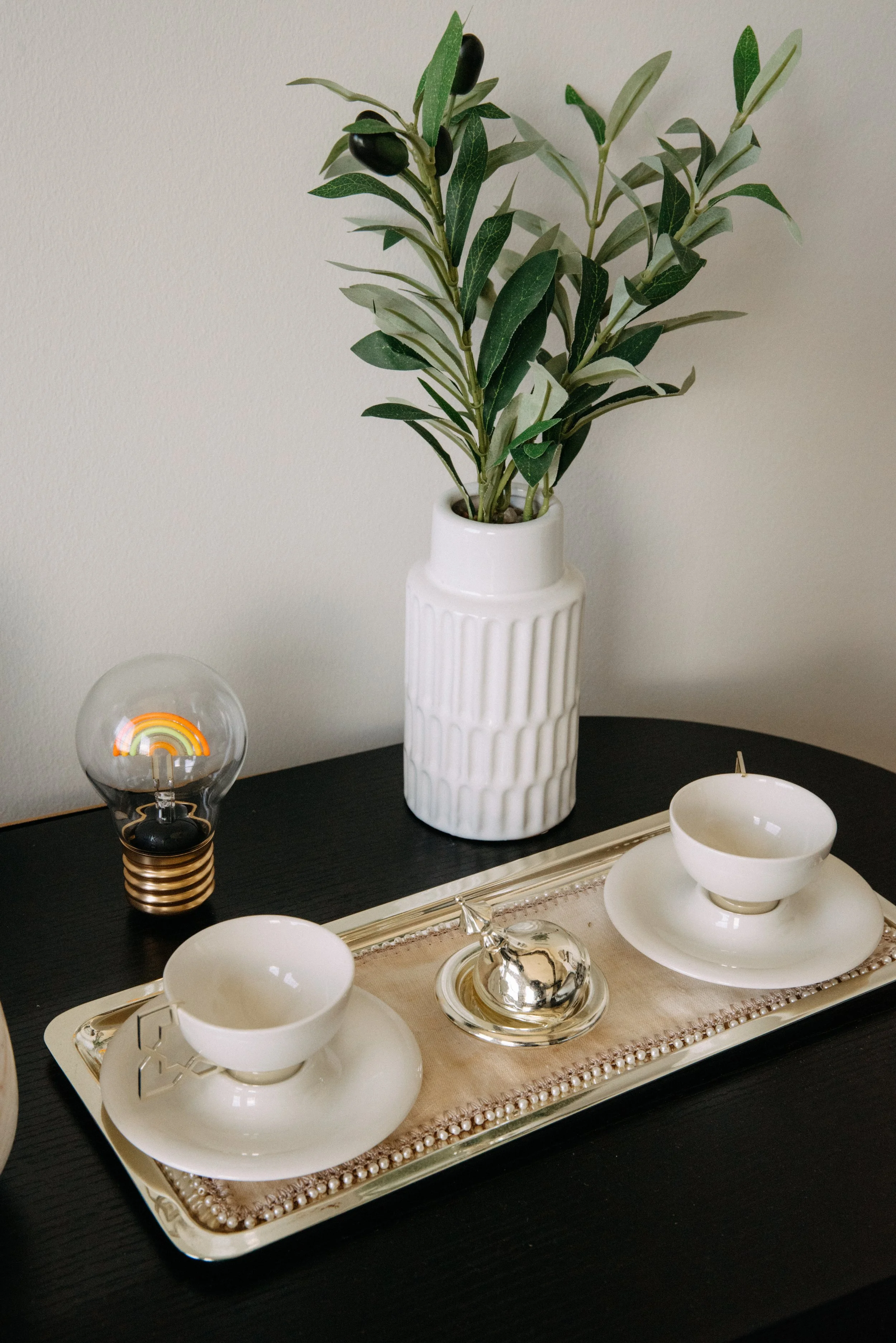 Decorative table setting with two white teacups and saucers, a silver ornament, a glass lamp with a rainbow design inside, a white vase with green foliage, and a tray with a beige runner on a dark table.