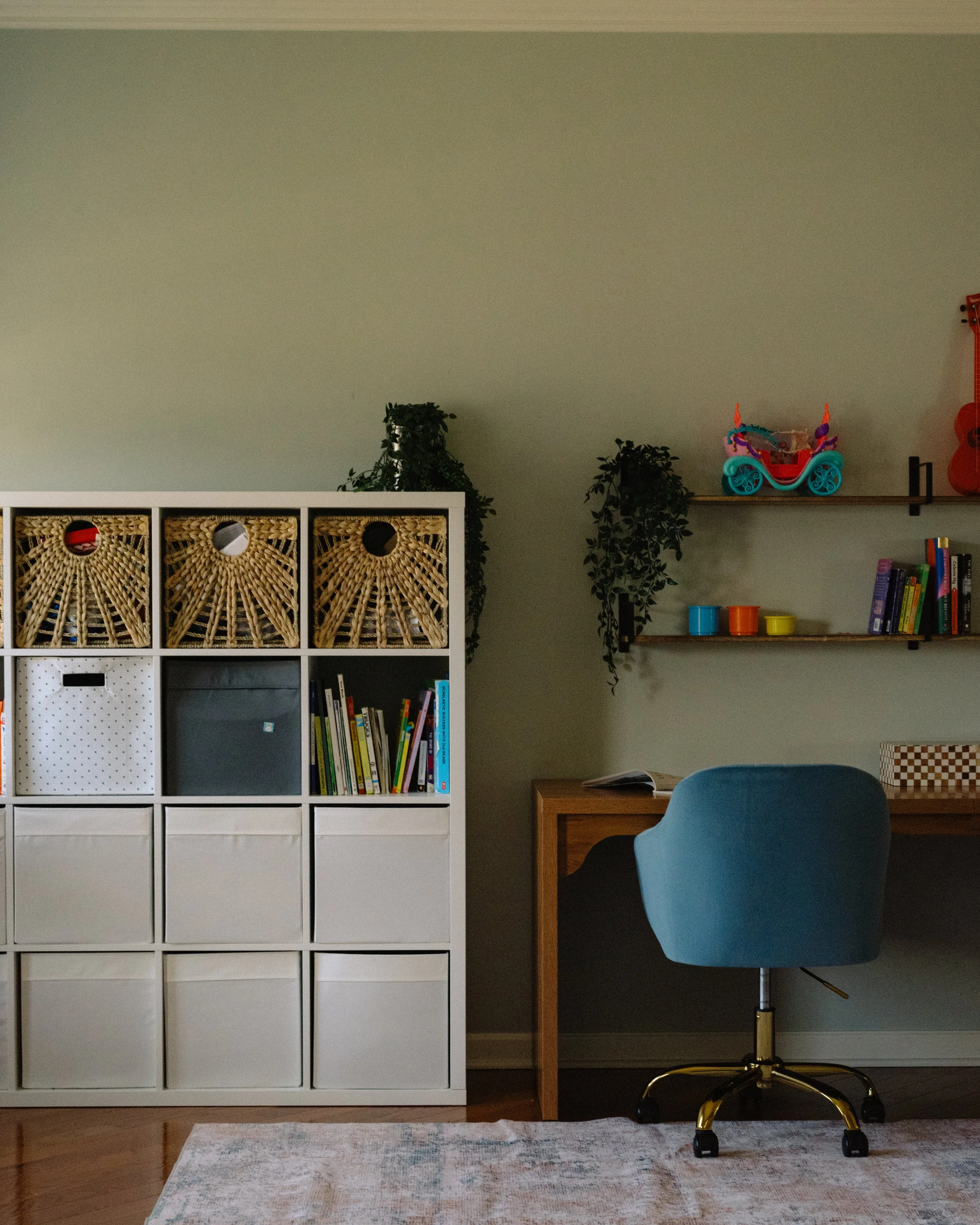 Home office with white storage cubby, bookshelf, blue office chair, and wall shelves with toys and books.