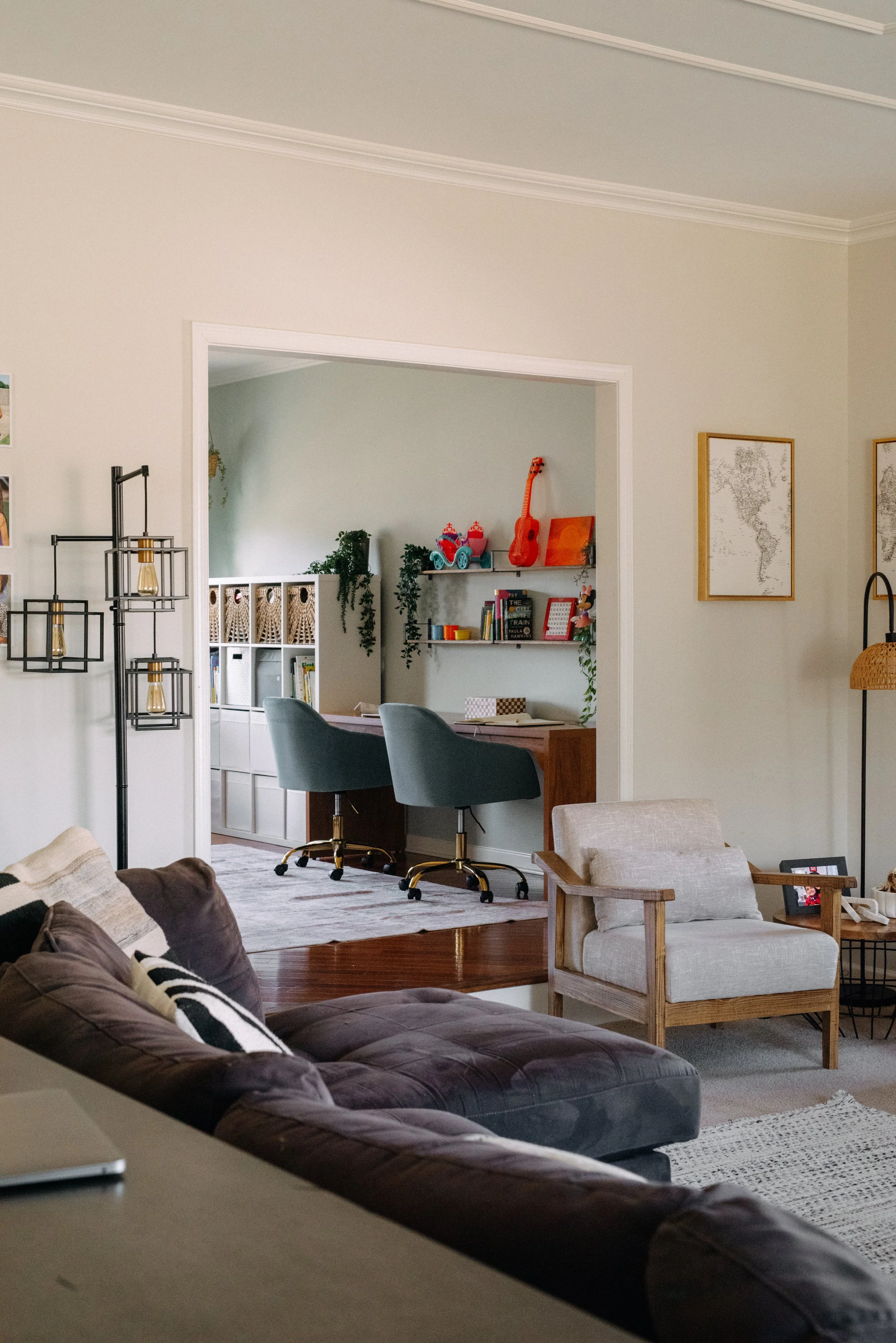 Living room with brown sofa, beige armchair, and a view into a workspace with two teal chairs and shelves with children's toys and decor.