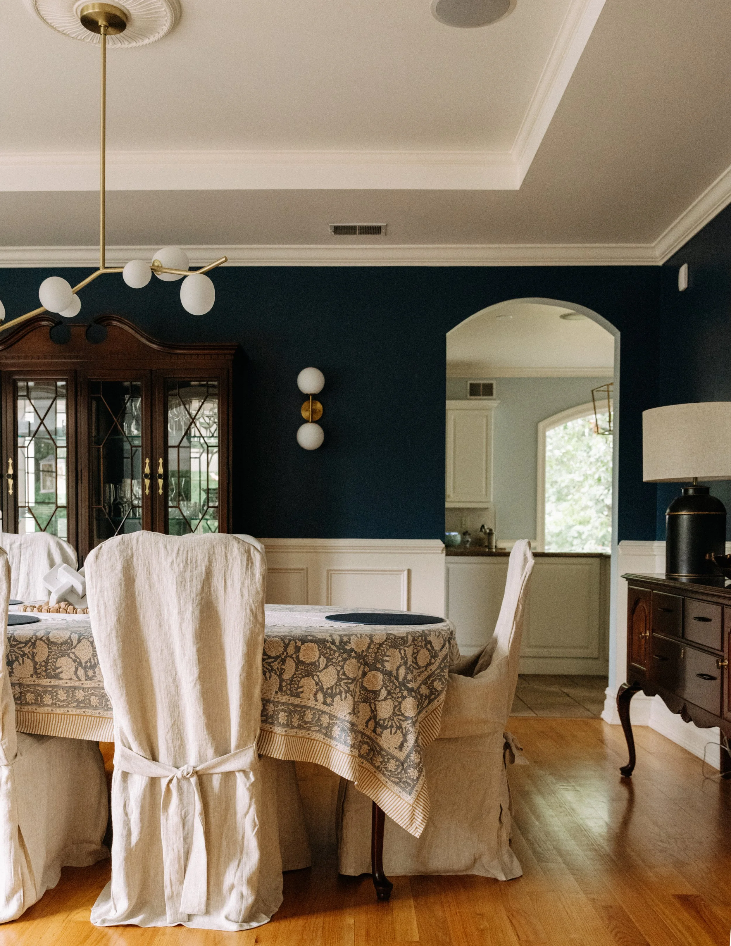 A dining room with a dark blue wall, cream chairs, a round table with a patterned tablecloth, a dark wooden china cabinet, a multi-bulb modern chandelier, a wall-mounted light fixture, and a sideboard with a lamp, with a view into a bright kitchen th