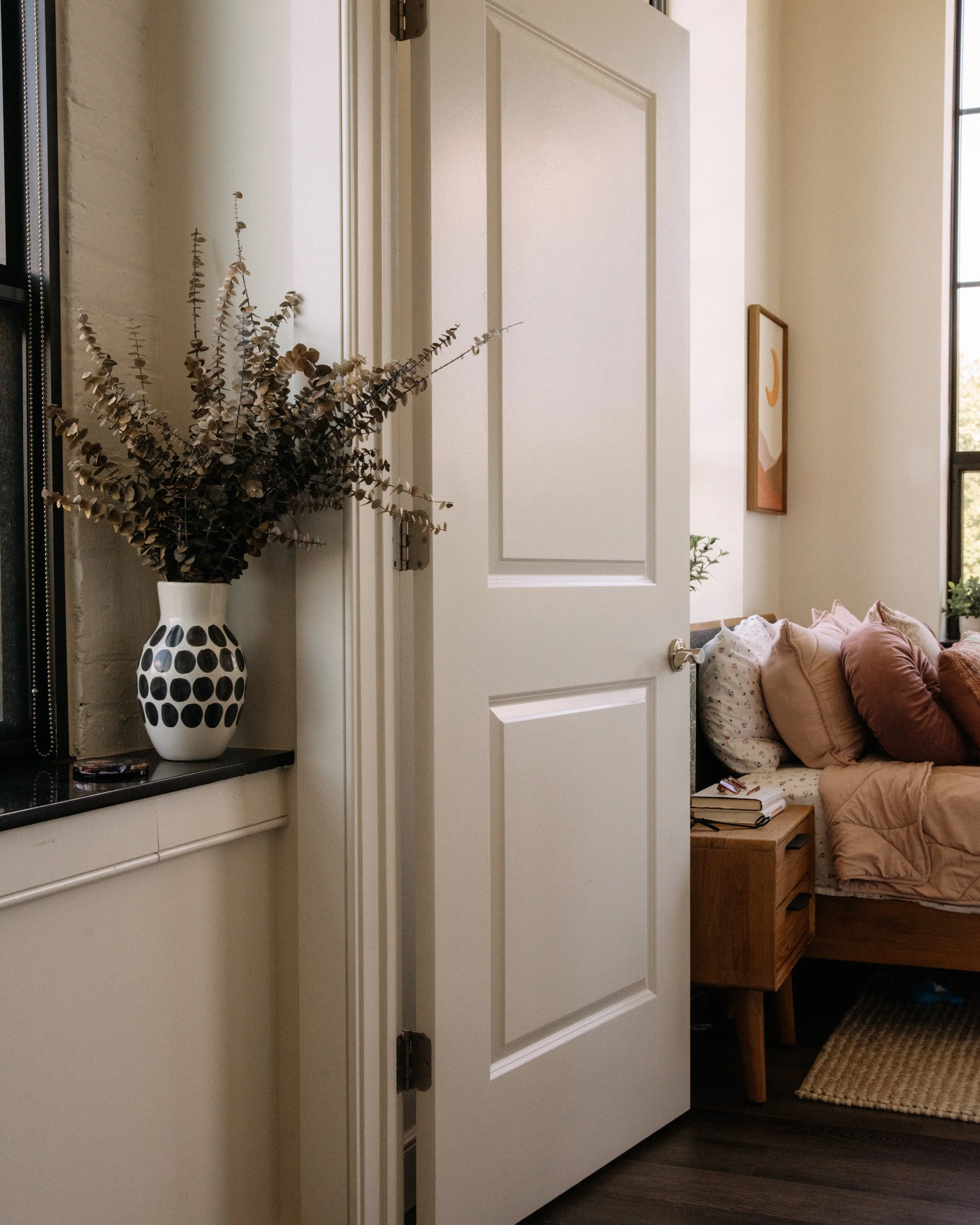 An open white door reveals a cozy living room with a sofa, plush pillows, a side table, and framed artwork on the wall, with sunlight streaming through a window.
