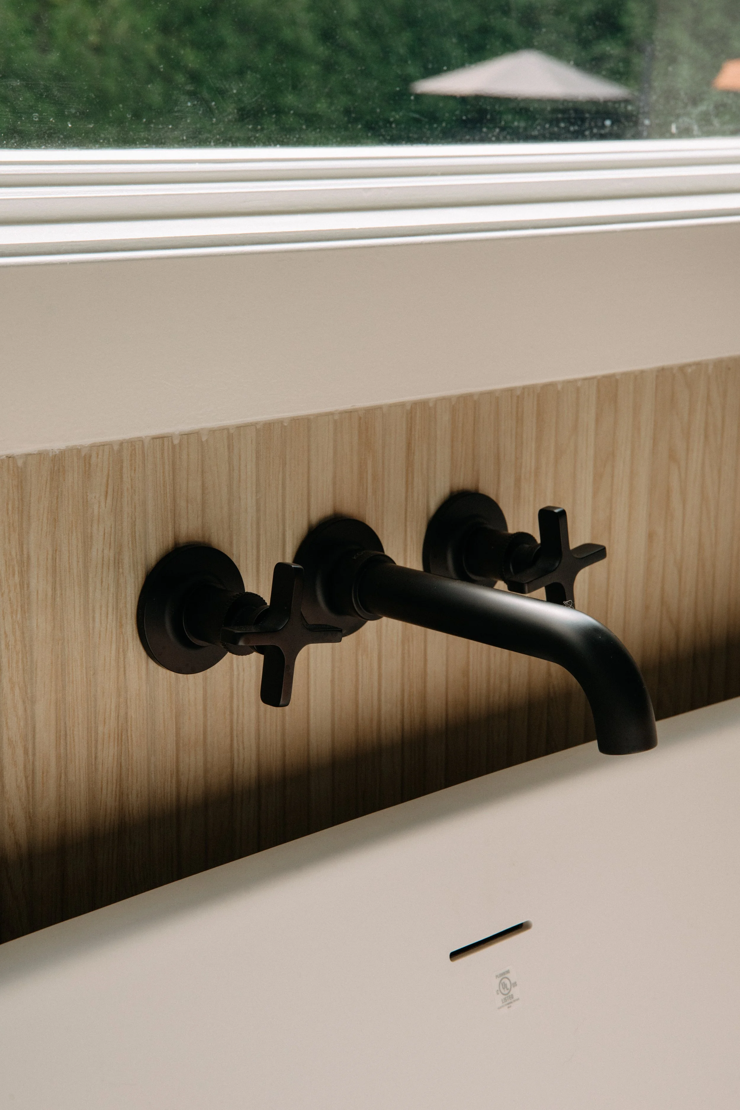 Close-up of black wall-mounted faucet with two handles above a white sink, against a wooden backsplash. Outside window with a blurred view of a patio umbrella visible.