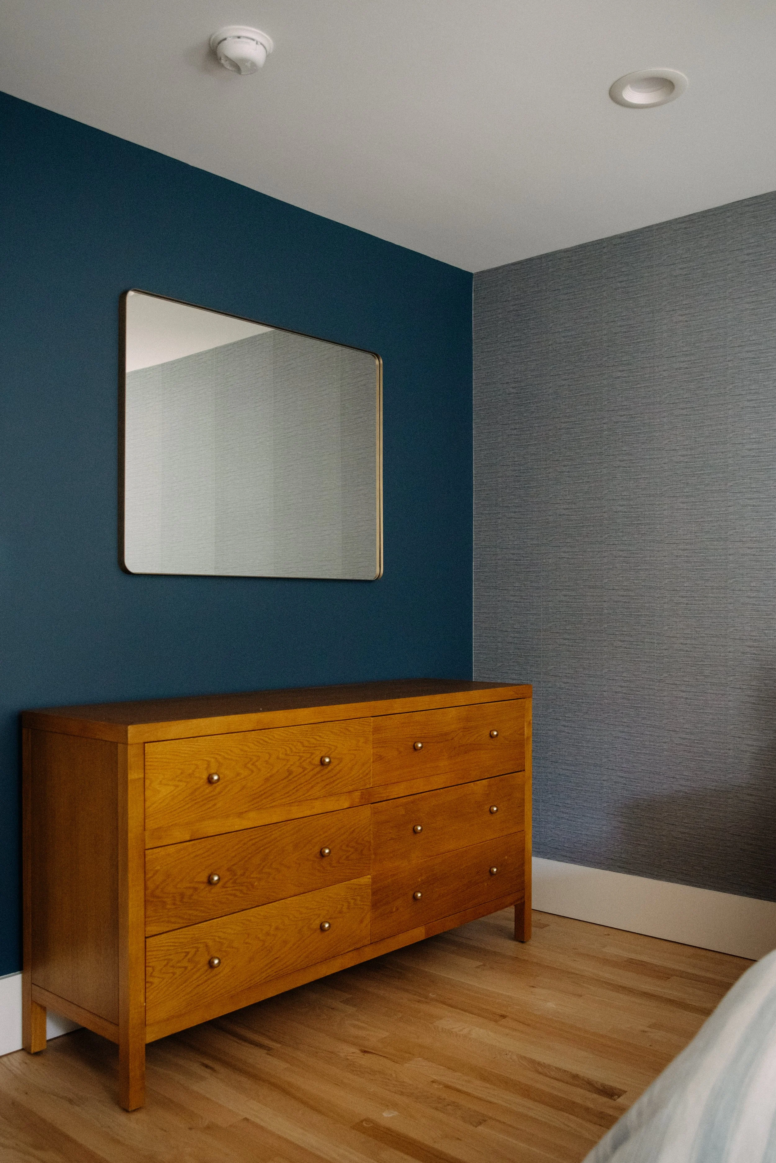 A wooden dresser with six drawers and round knobs, placed against a dark blue wall with a mirror above it, in a room with gray textured wall and hardwood flooring.