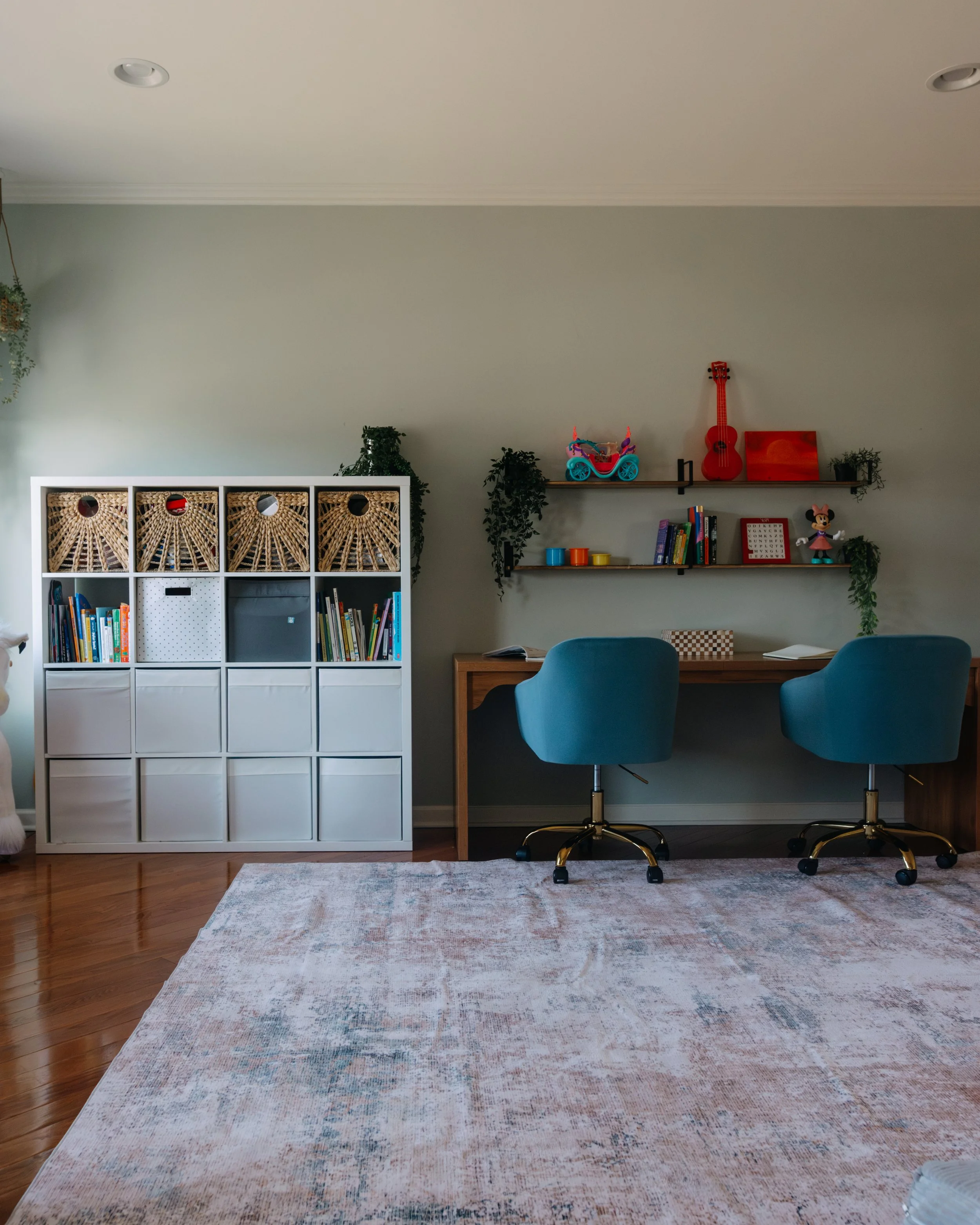 A home office with two blue chairs in front of a wooden desk, shelves with toys and books, a white storage unit, and a beige rug on a wooden floor.