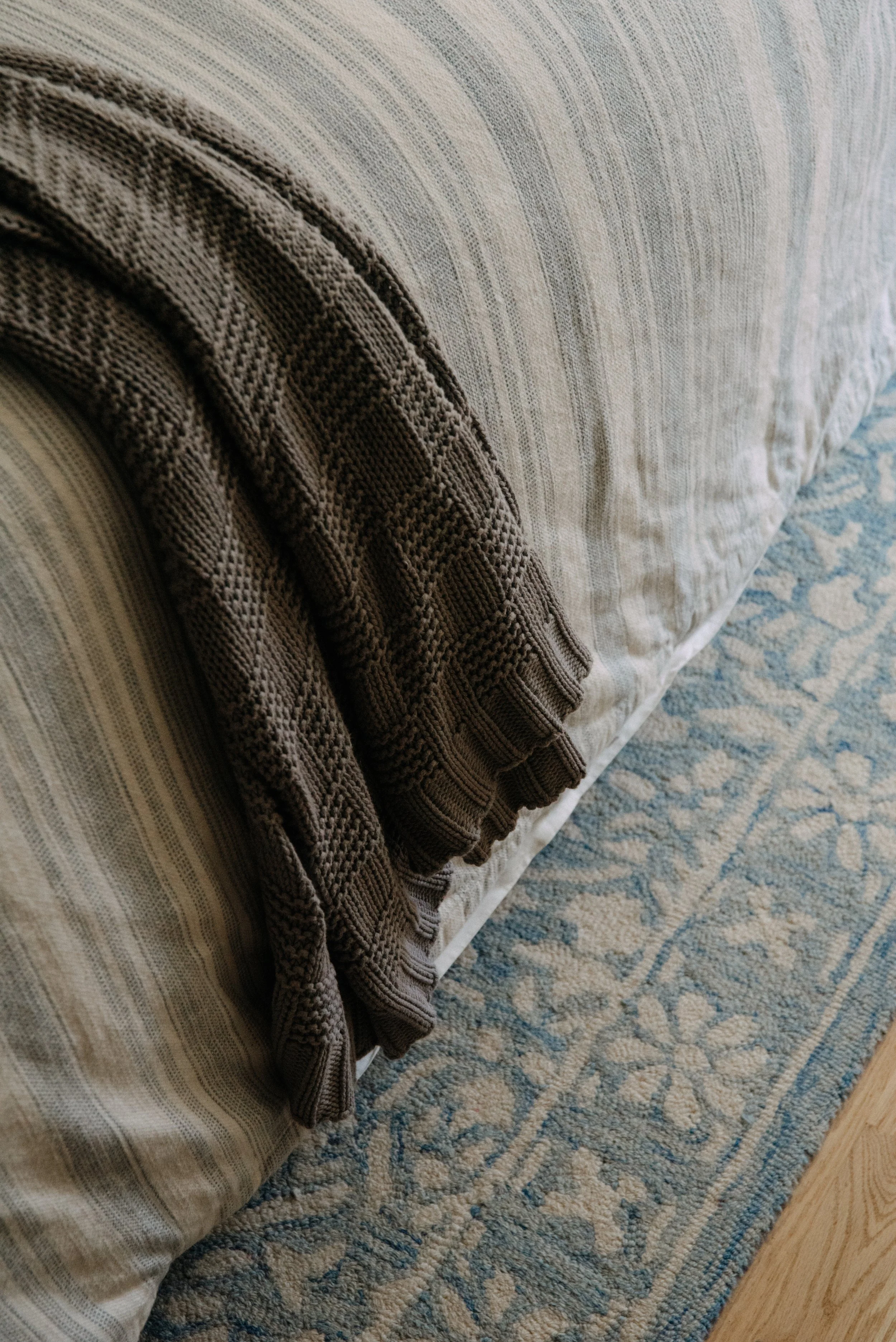 Folded knit blanket on a bed with a beige and cream striped duvet, sitting on a patterned blue and beige rug.