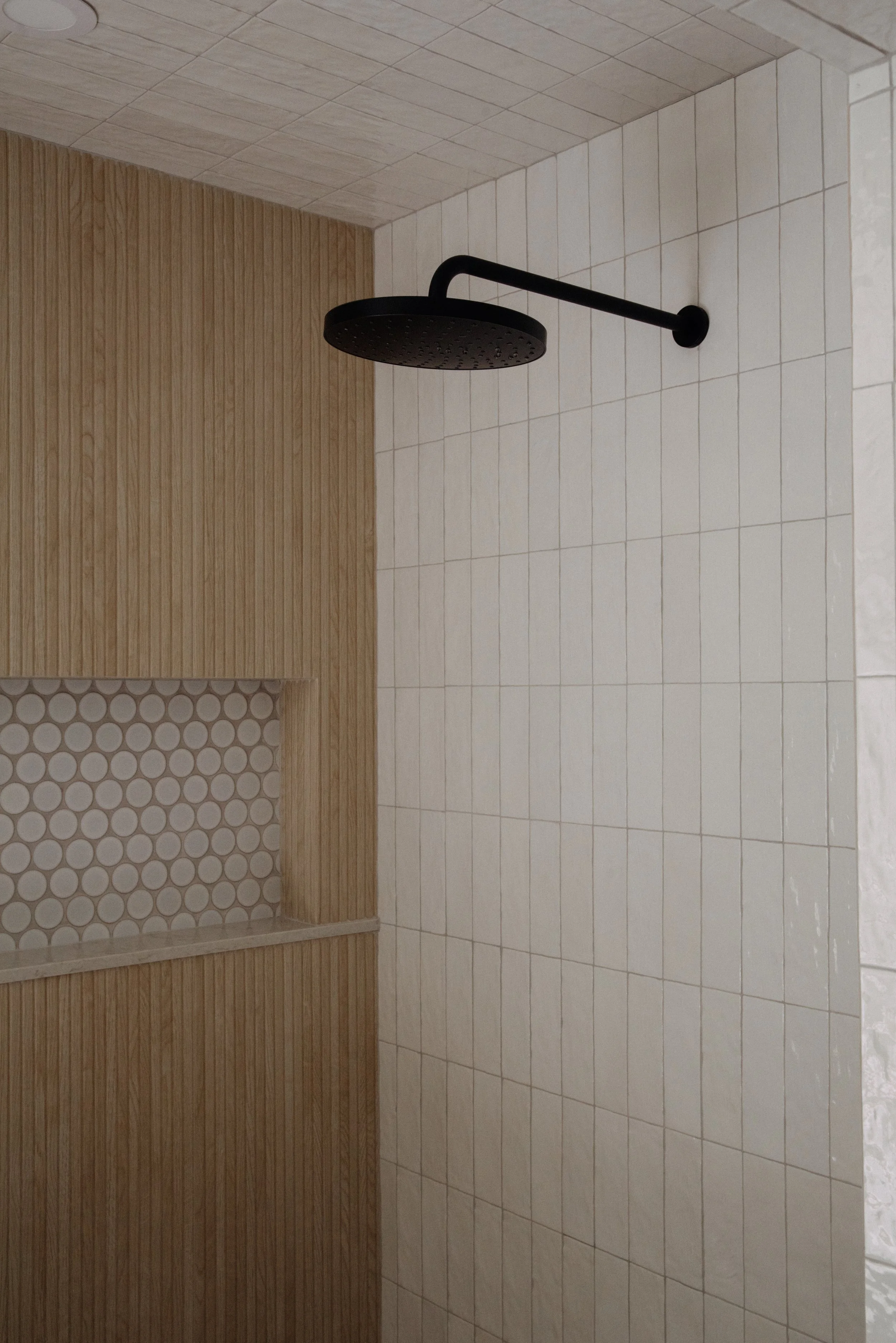 A shower area with sepia tones, featuring a black showerhead, a wooden accent wall, and light-colored ceramic tiles, including a small built-in shelf with circular tile pattern.