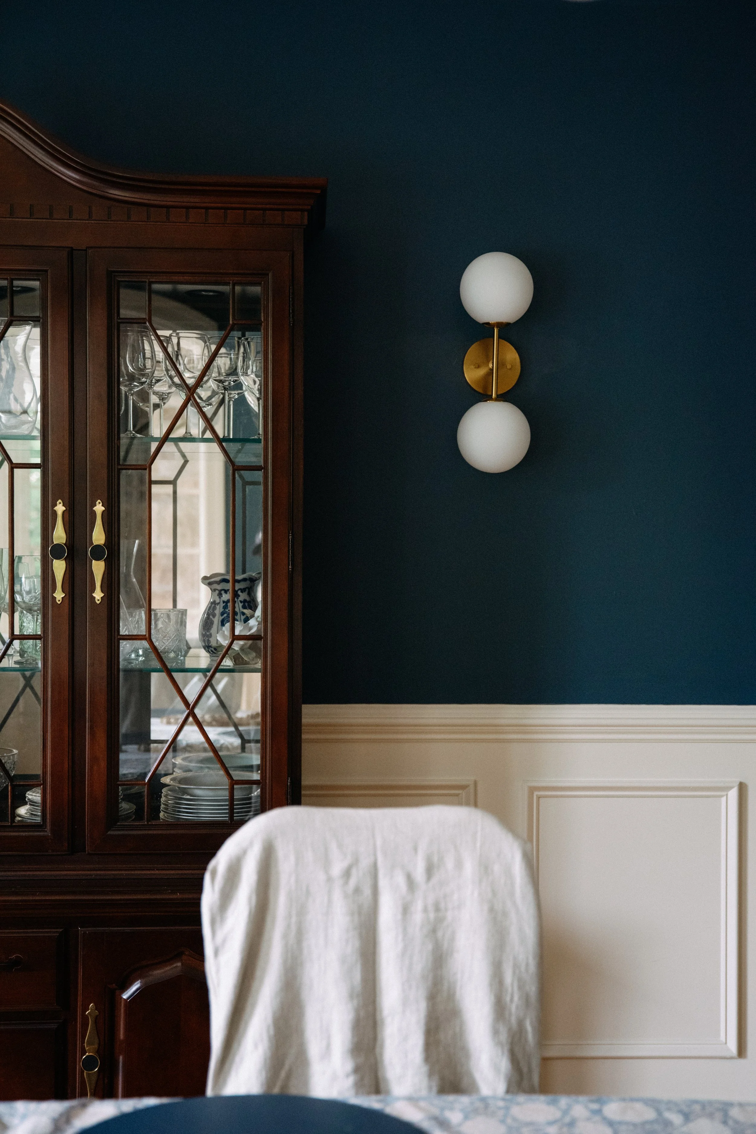 A side view of a dark wooden china cabinet with glass doors containing glassware and dishes, and a modern wall sconce with two white globes on a dark blue wall behind a white chair with a cloth cover