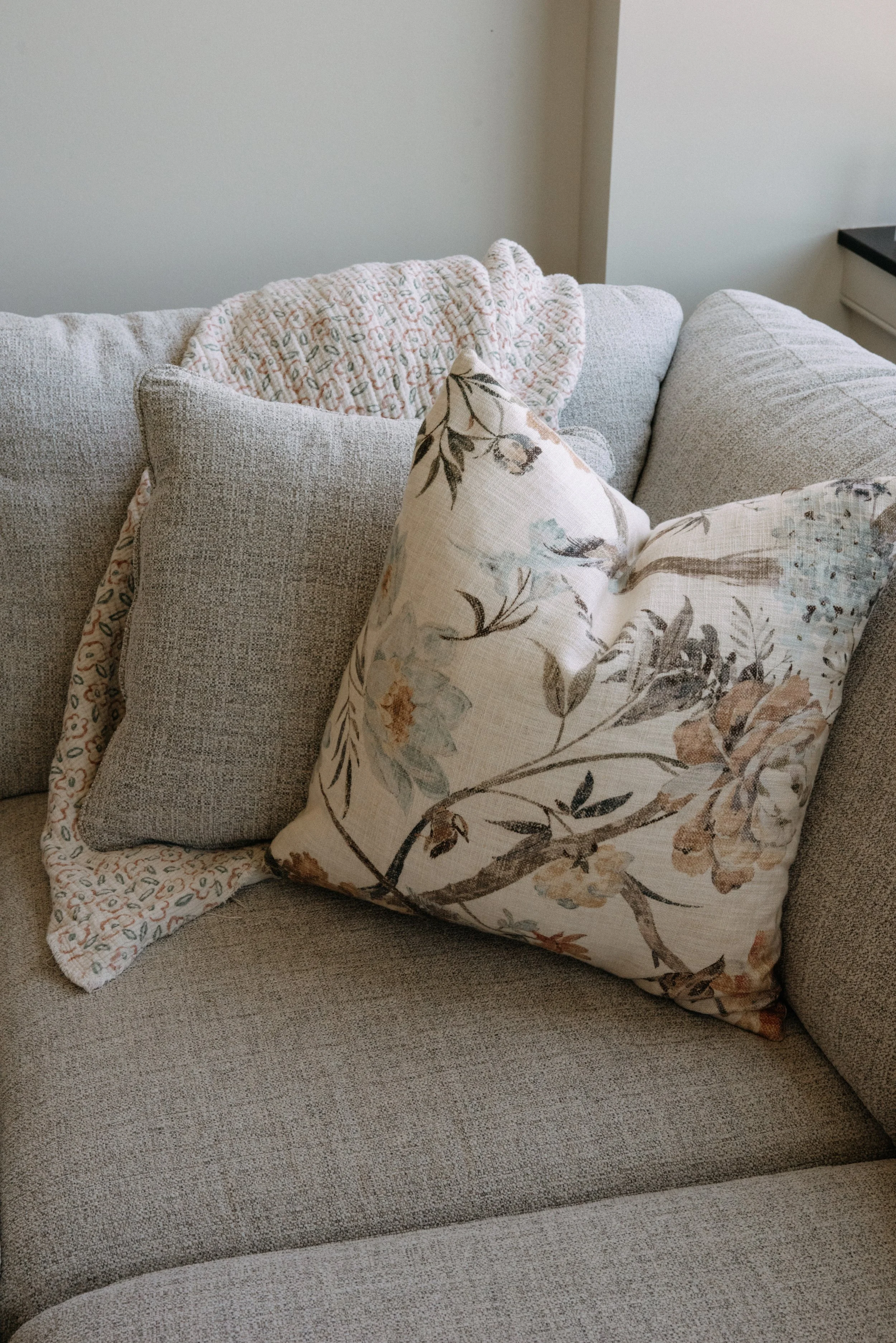 A beige fabric sofa with multiple throw pillows, including a floral patterned pillow and a small, textured pillow, with a quilted blanket draped over the back.