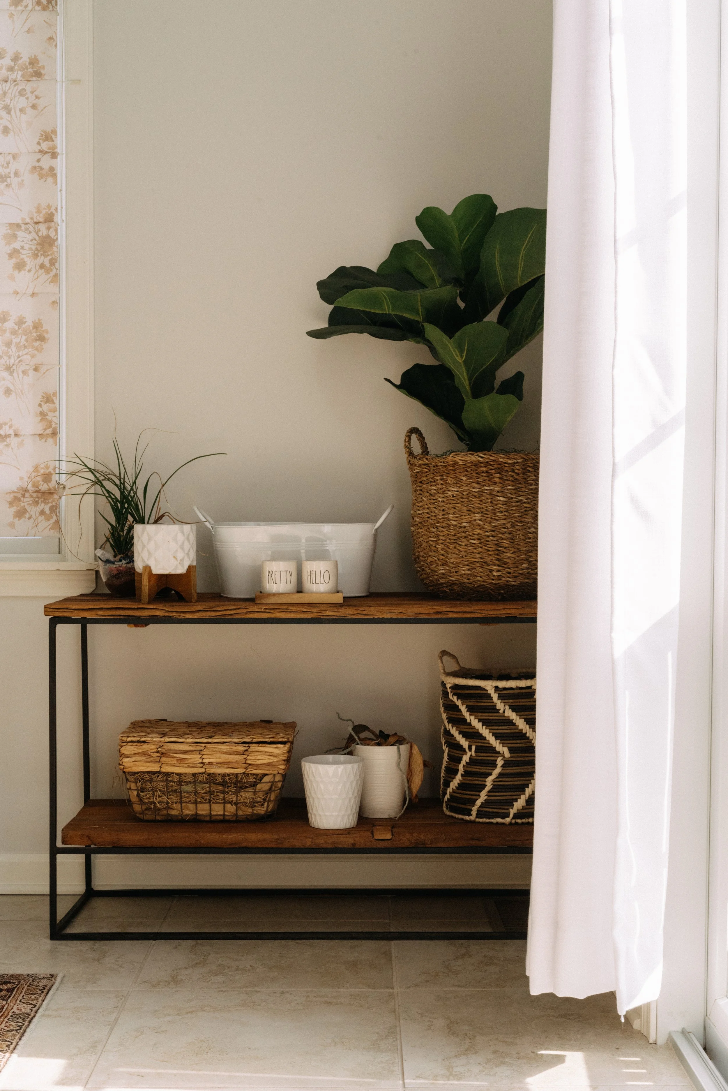 A wooden and metal console table with plants, decorative containers, and baskets, situated next to white curtains and a window with floral blinds.