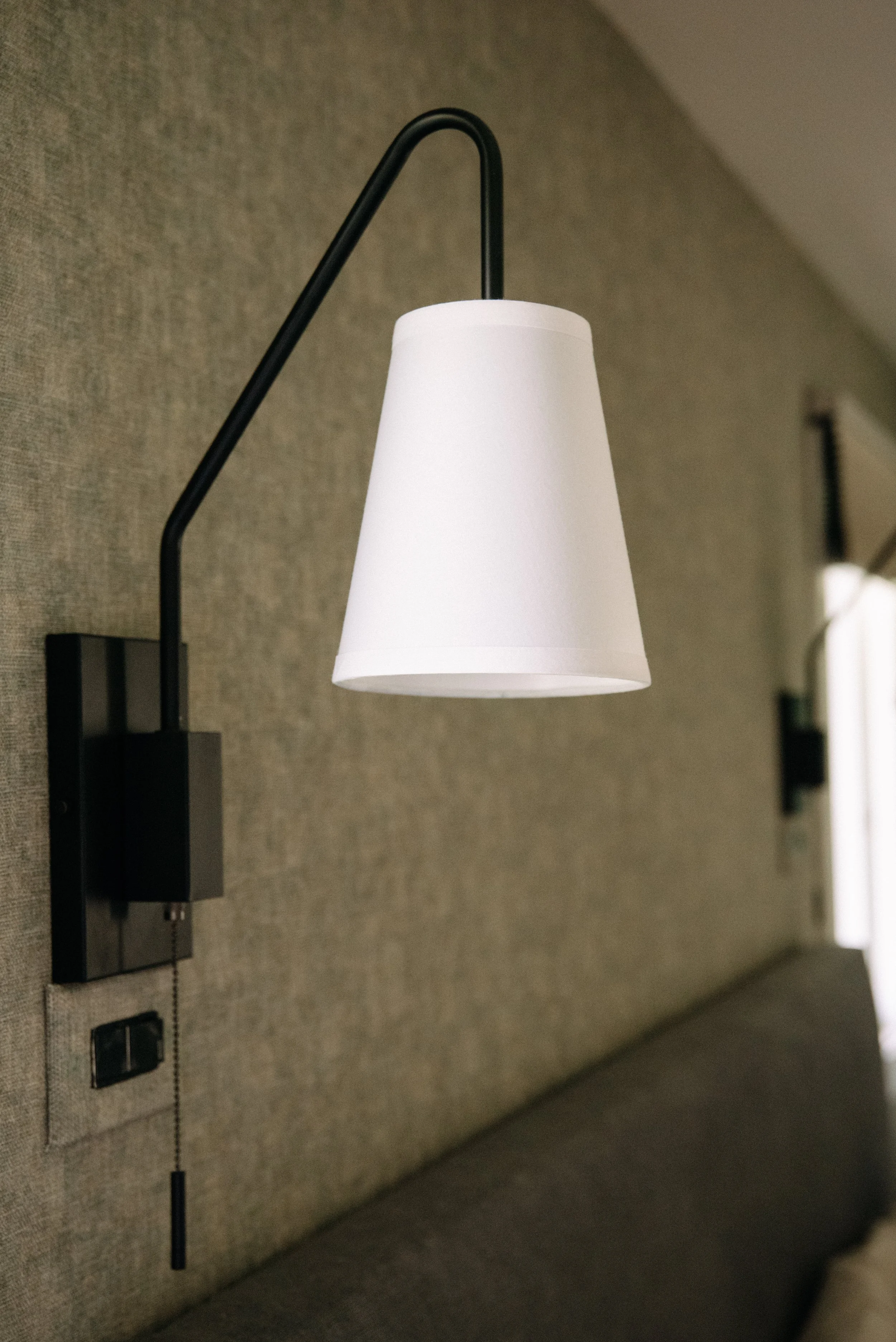 Wall-mounted black and white adjustable reading lamp with a conical white lampshade, installed on a textured beige wall above a bed.