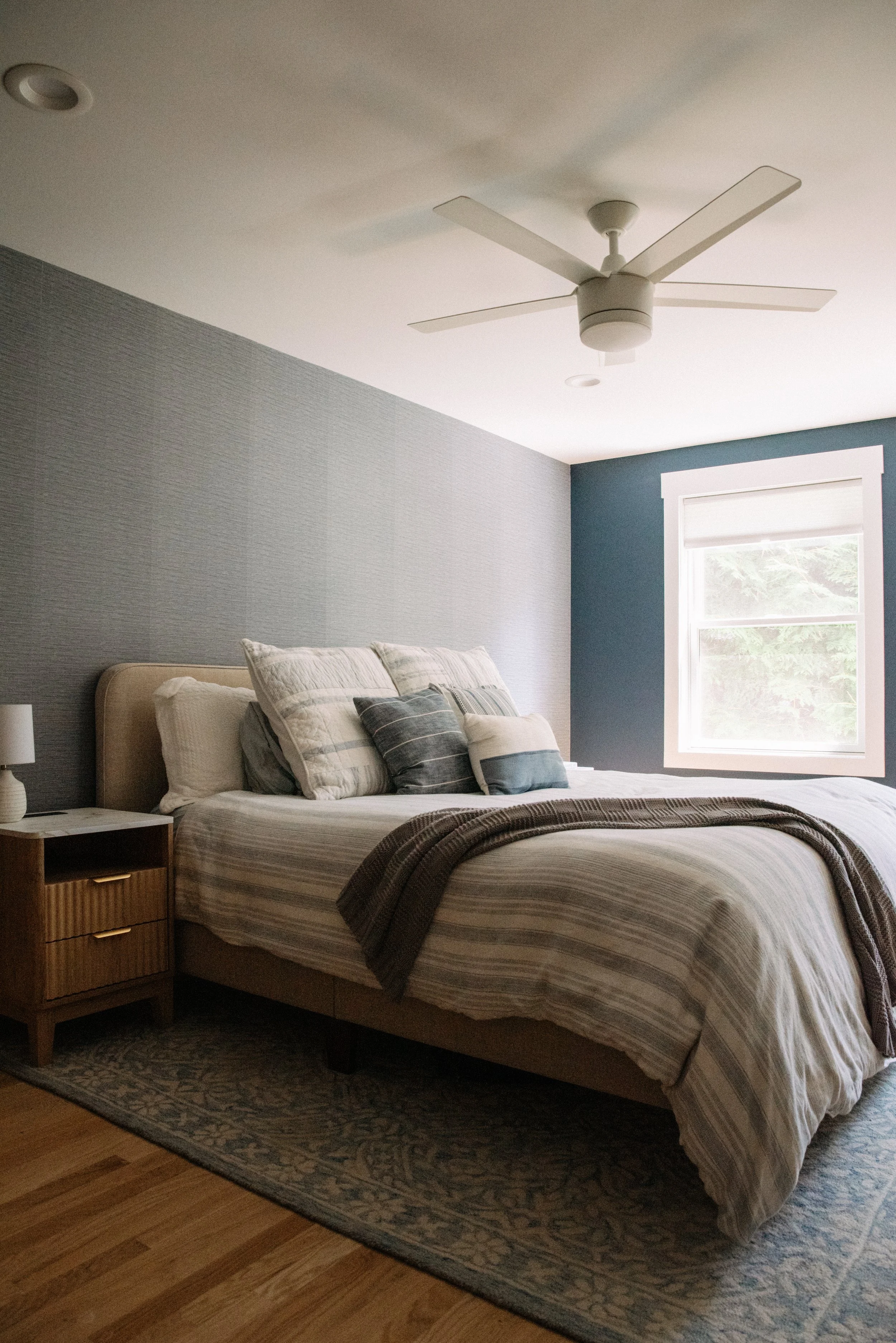 A bedroom with a bed, pillows, a nightstand with a lamp, a ceiling fan, and a window with white trim letting in natural light.