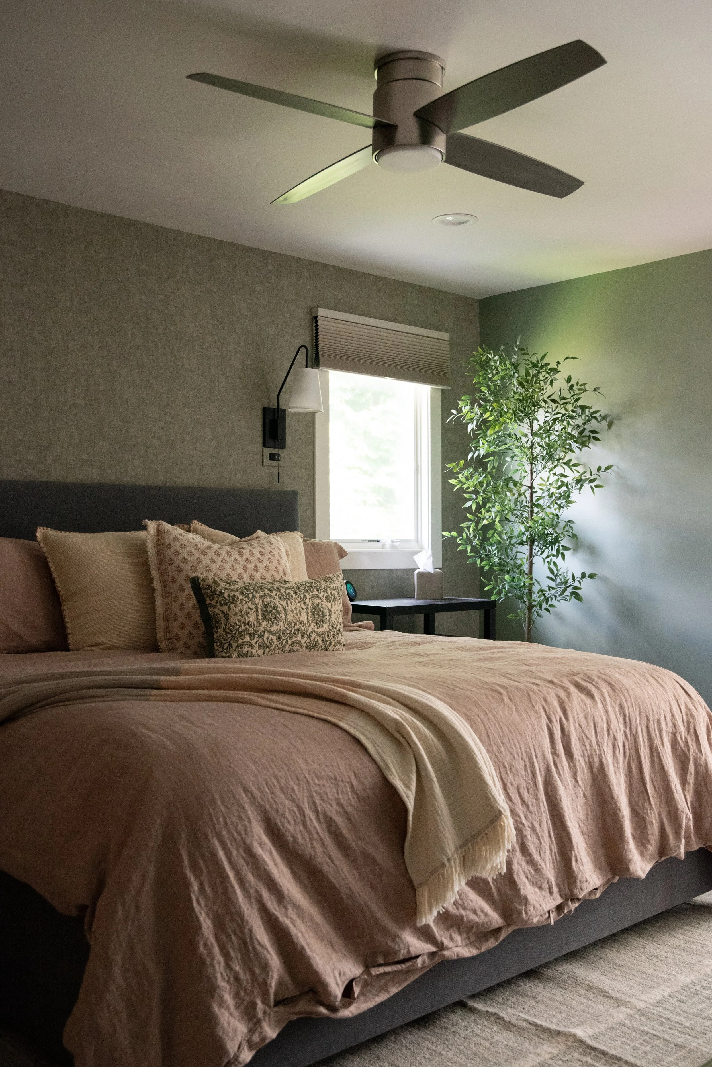 A cozy bedroom with a bed made with beige and pink bedding, a pillow with a floral pattern, a small black side table with a tissue box and a clock, a window with a beige blind, a tall green plant, and a ceiling fan.