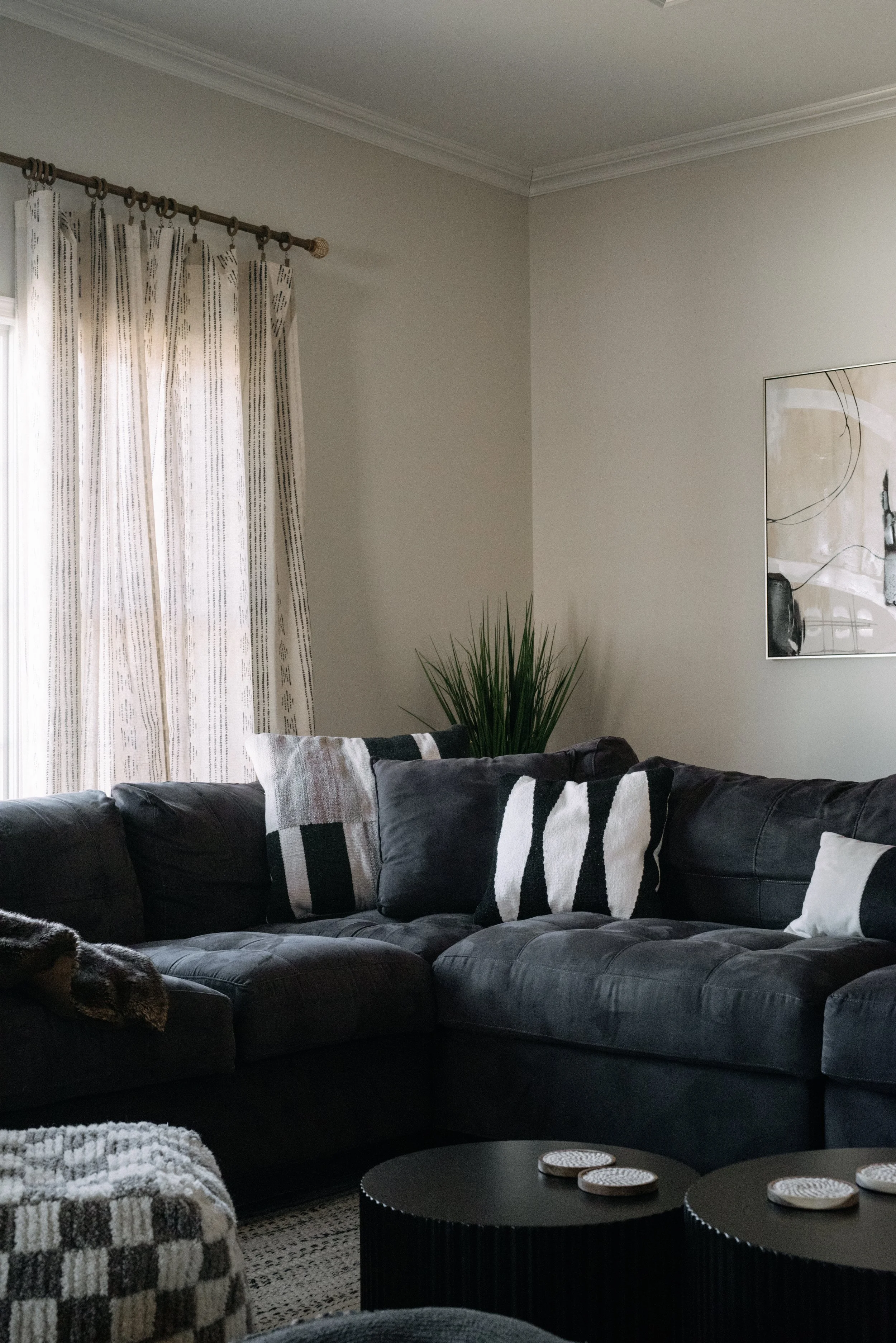 Living room with a black sectional sofa, patterned pillows, a tall plant in the corner, curtains on a window, a wall art piece, and two black coffee tables with coasters.