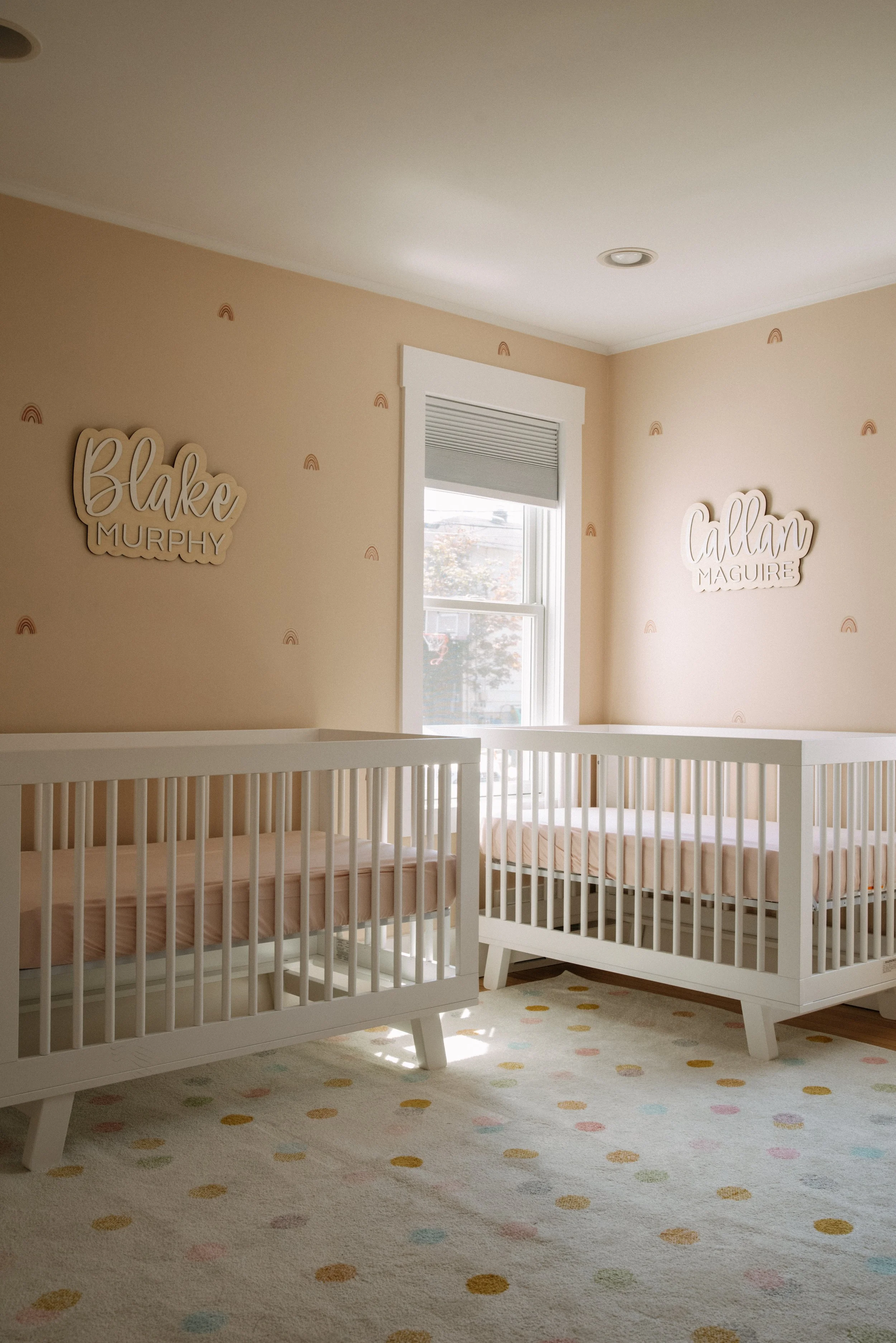 A nursery with two white cribs, one on each side, near a window with a partially closed white blind. The walls are painted a light beige with small rainbow decals. There are wooden name signs on the walls: "Blake Murphy" on the left and "Callan Magui